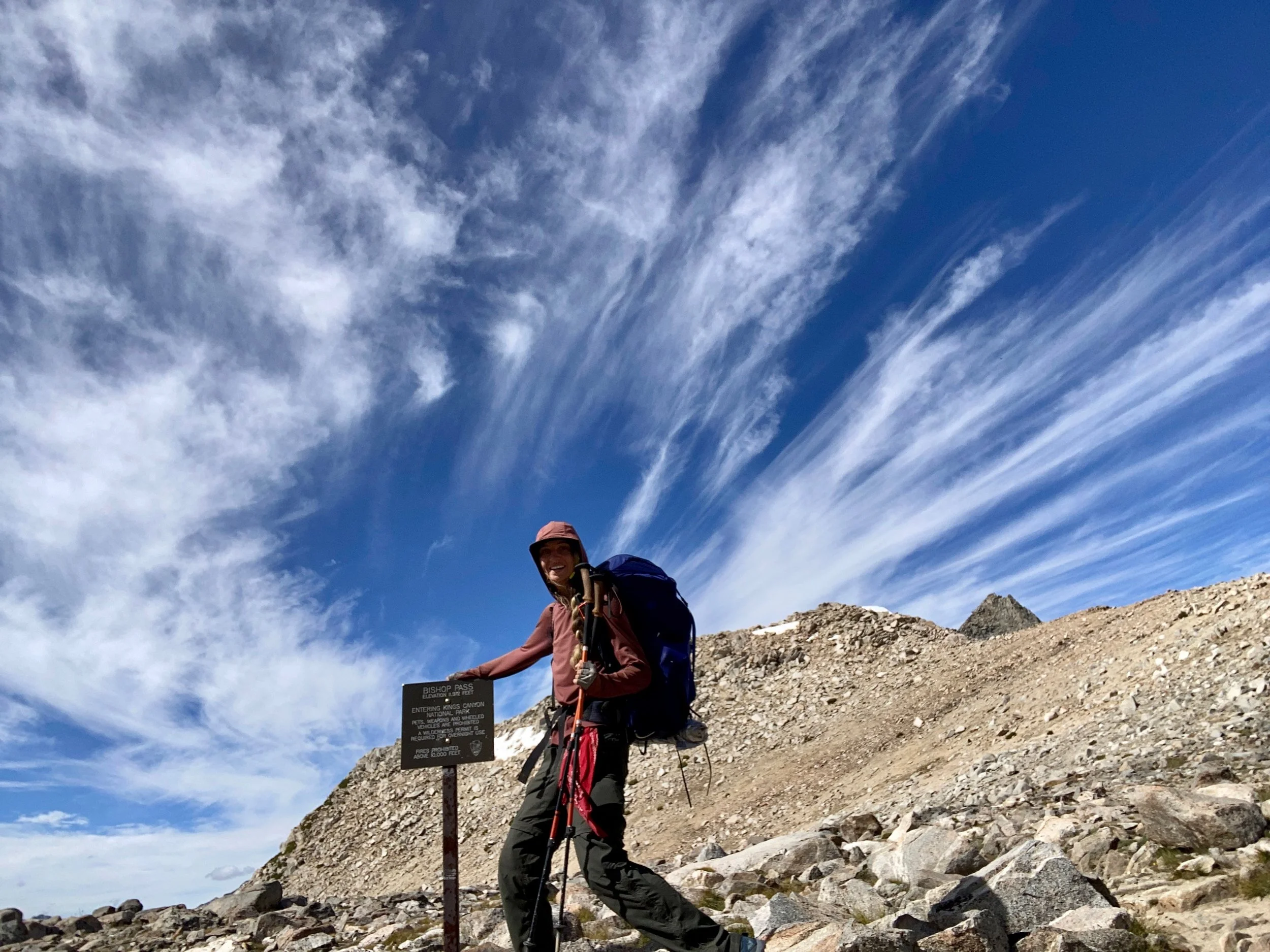 A woman in outdoor hiking gear, holding trekking poles, standing on a rocky mountain trail under a partly cloudy sky with wispy clouds. There is a signpost next to her with information about the trail and park regulations.
