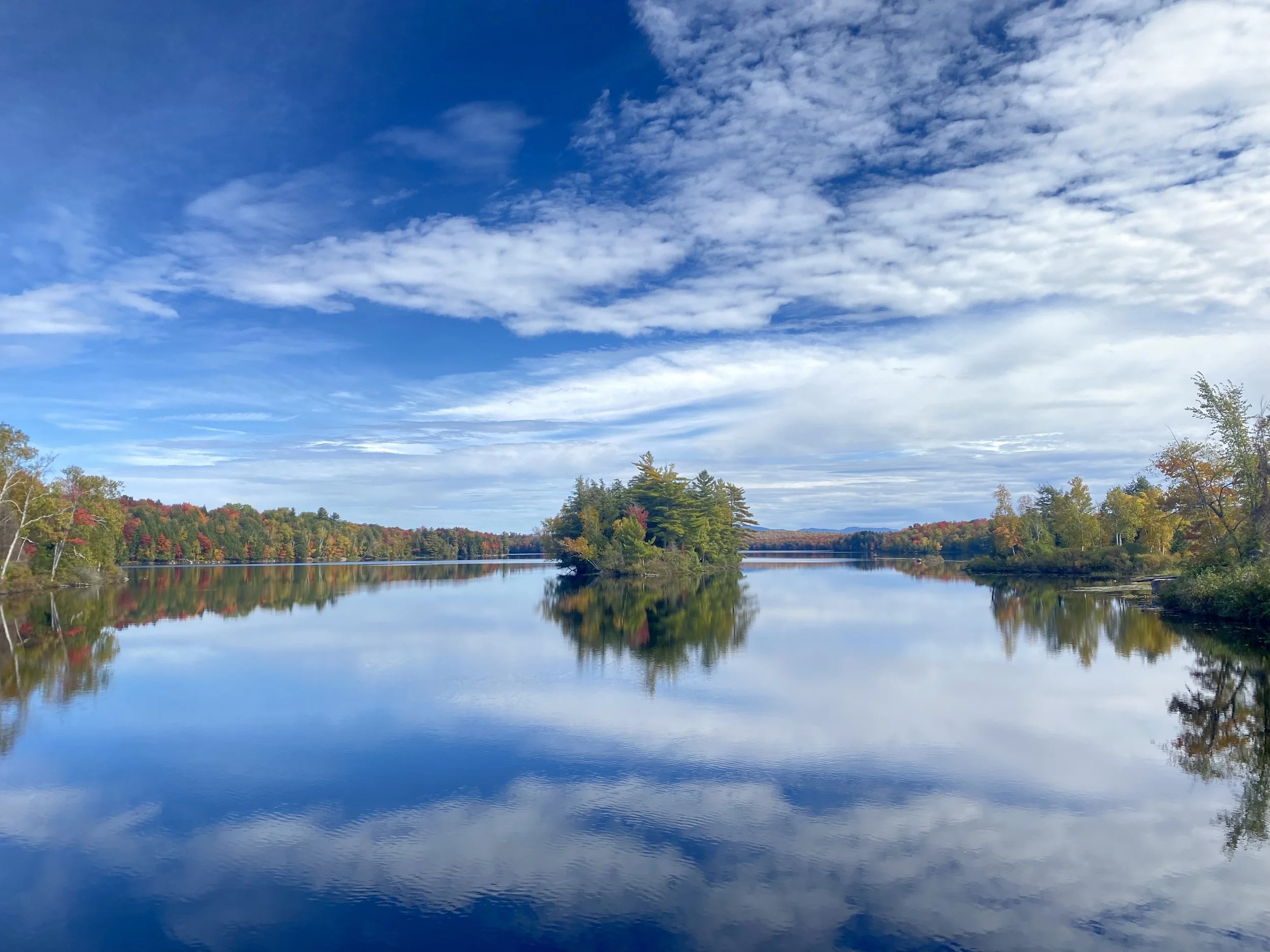 A calm lake reflecting a colorful autumn forest and a partly cloudy blue sky.