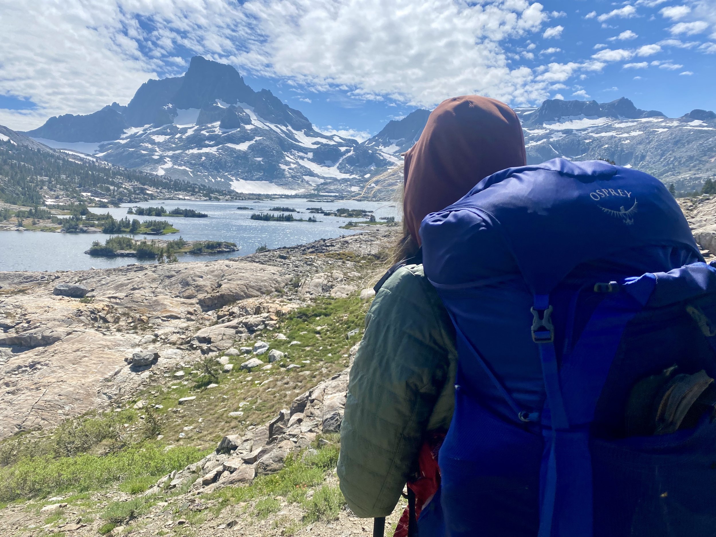A person with a brown hooded jacket and a blue backpack sitting on rocks, overlooking a mountain landscape with snow-capped mountains, a lake, and a partly cloudy sky.