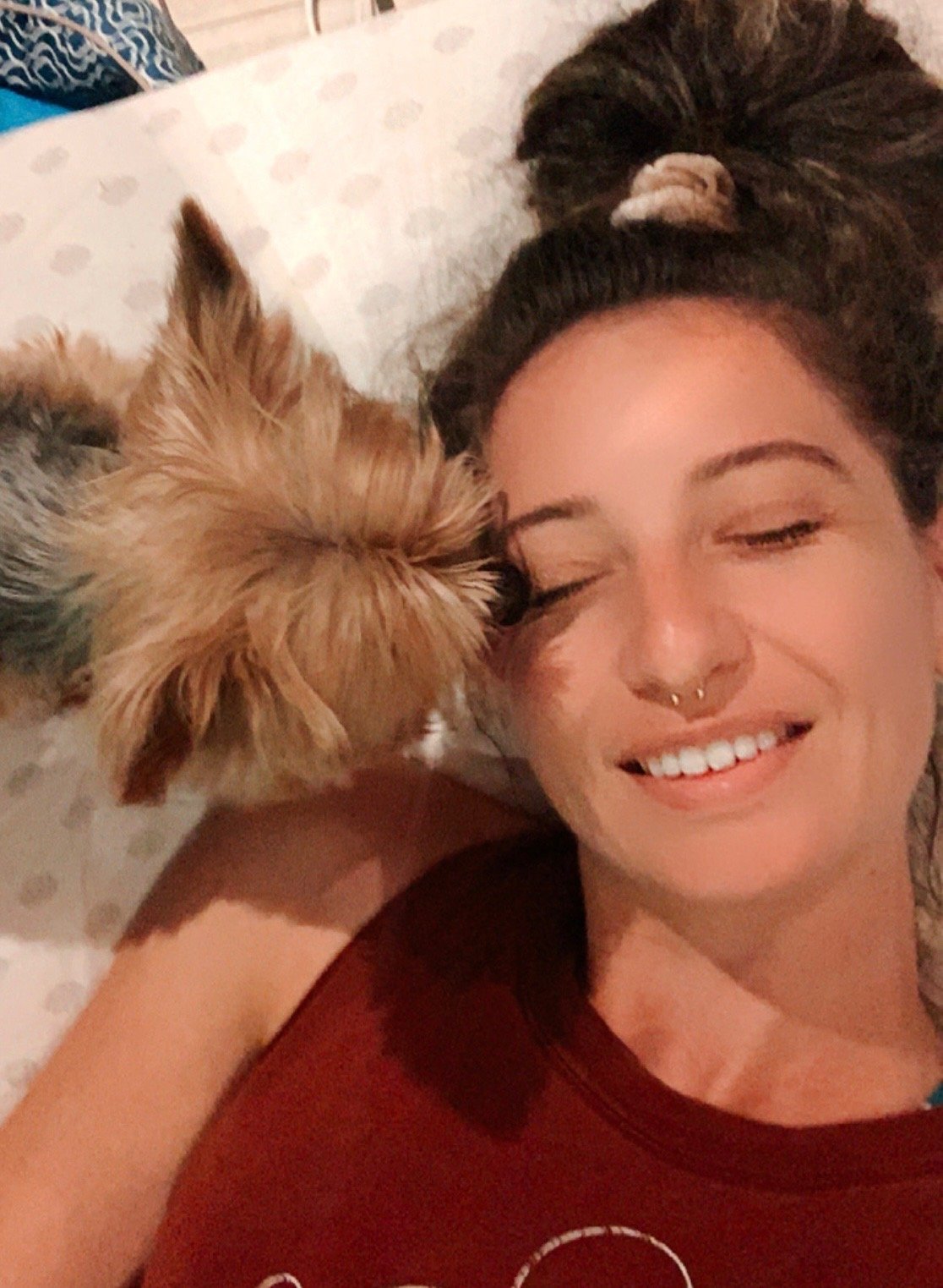 A young woman with dark hair in a messy bun, smiling with closed eyes, lying down next to a small, fluffy dog with brown and gray fur, also lying down with its face close to her.