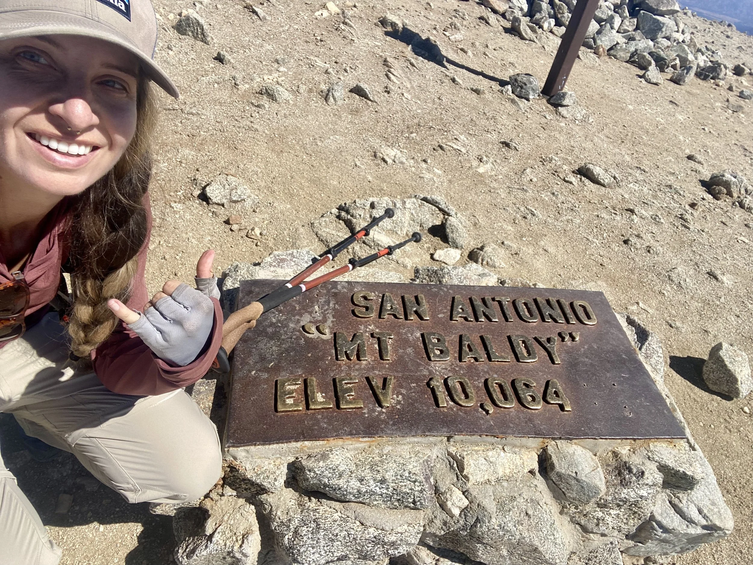 A smiling woman with a tan hat, glasses, and a nose piercing taking a selfie on Mount Baldy, California, standing next to a metal marker with engraved text, surrounded by rocky terrain.