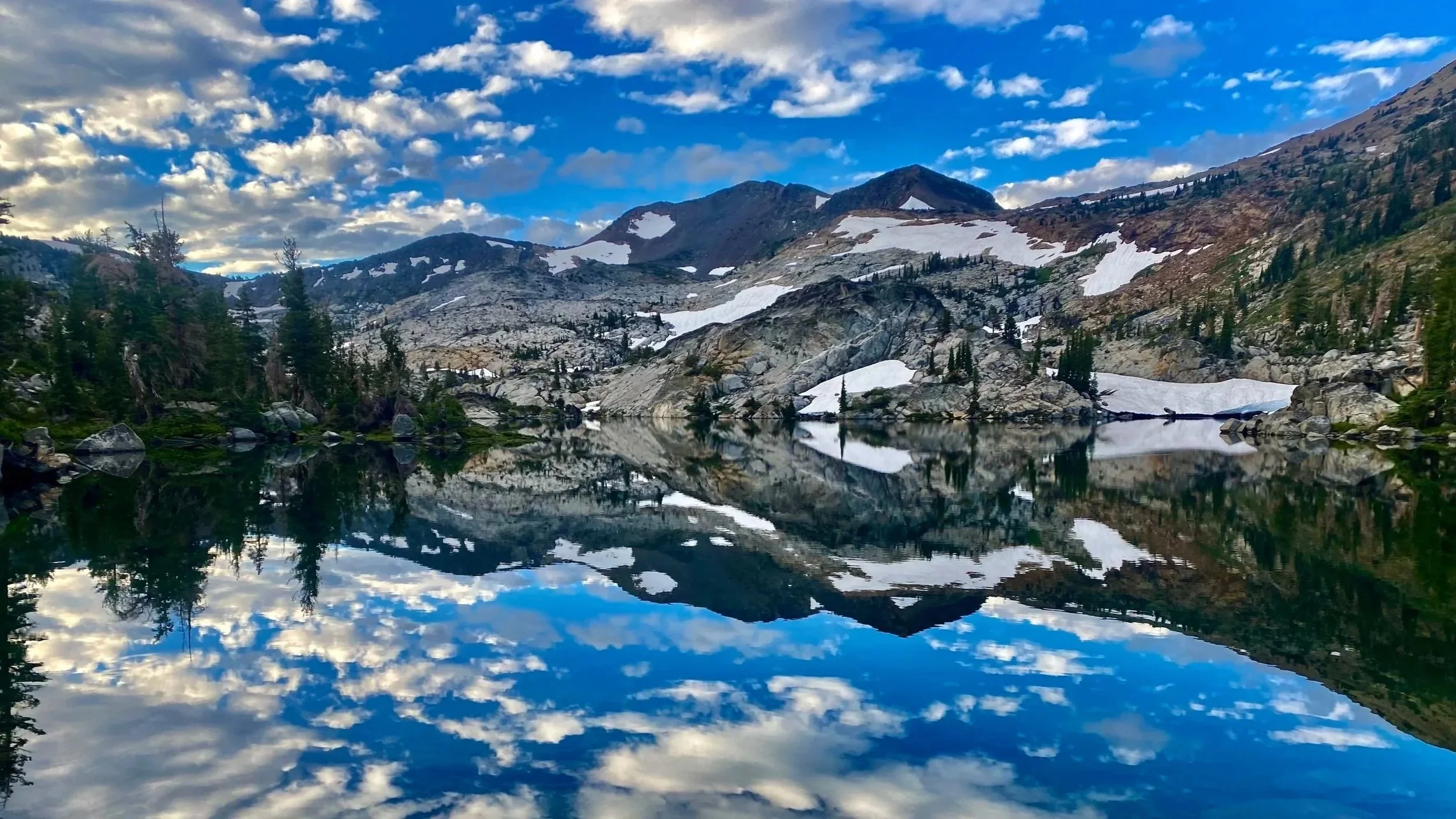 A mountain landscape features a clear lake reflecting snow-capped mountains, pine trees, and a partly cloudy sky.