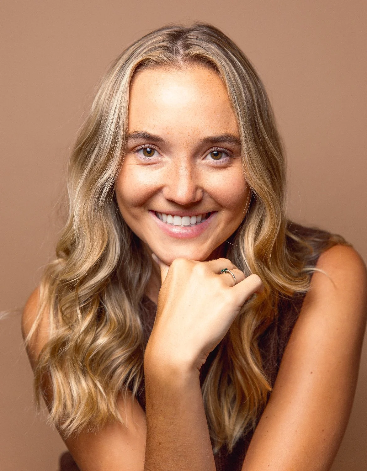 Smiling young woman with long wavy blonde hair resting her chin on her hand against a beige background.