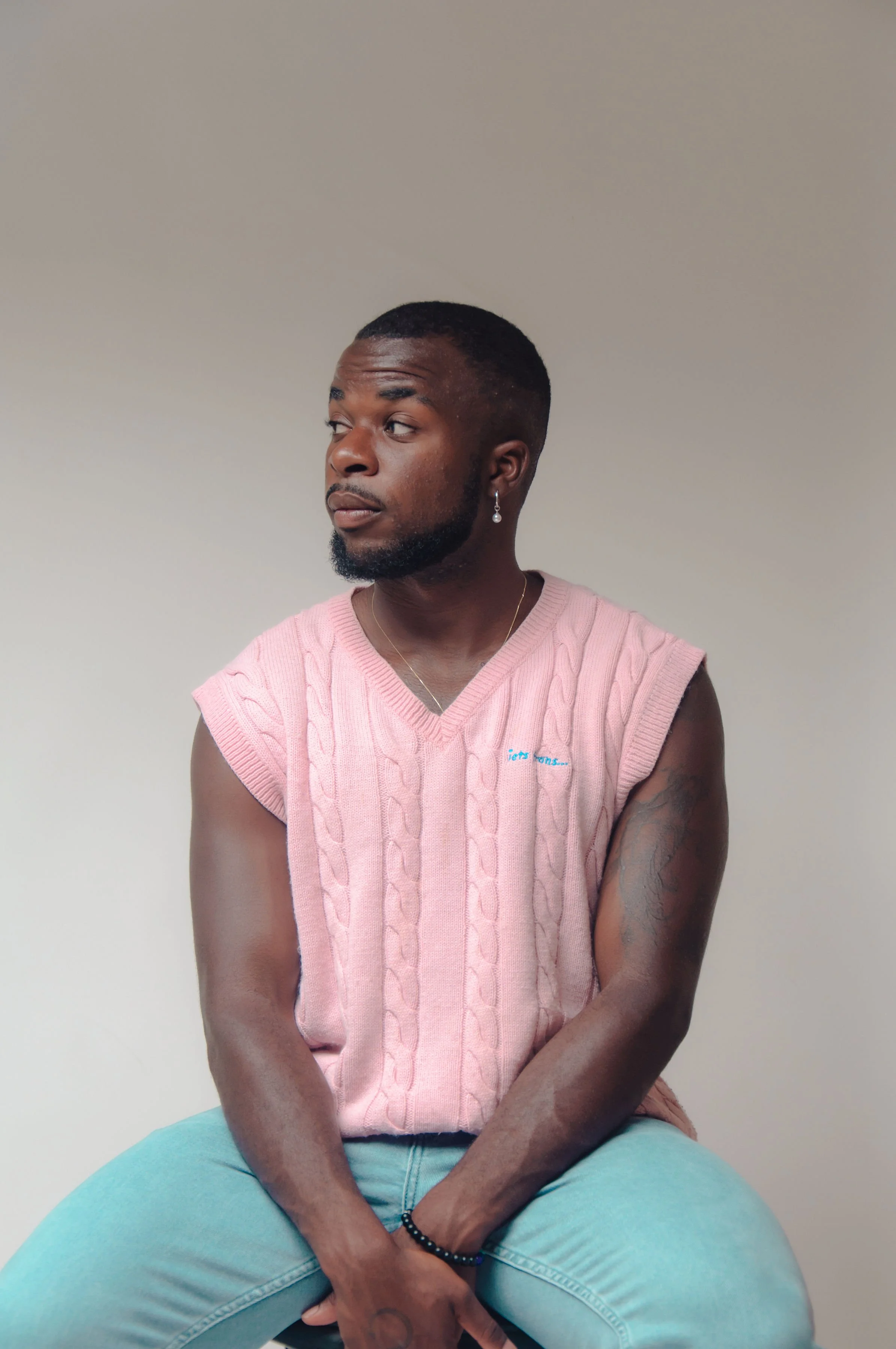 A man with dark skin, short hair, and a beard, wearing a pink sleeveless knitted sweater and light blue jeans, sitting against a plain light-colored background, looking to the side.