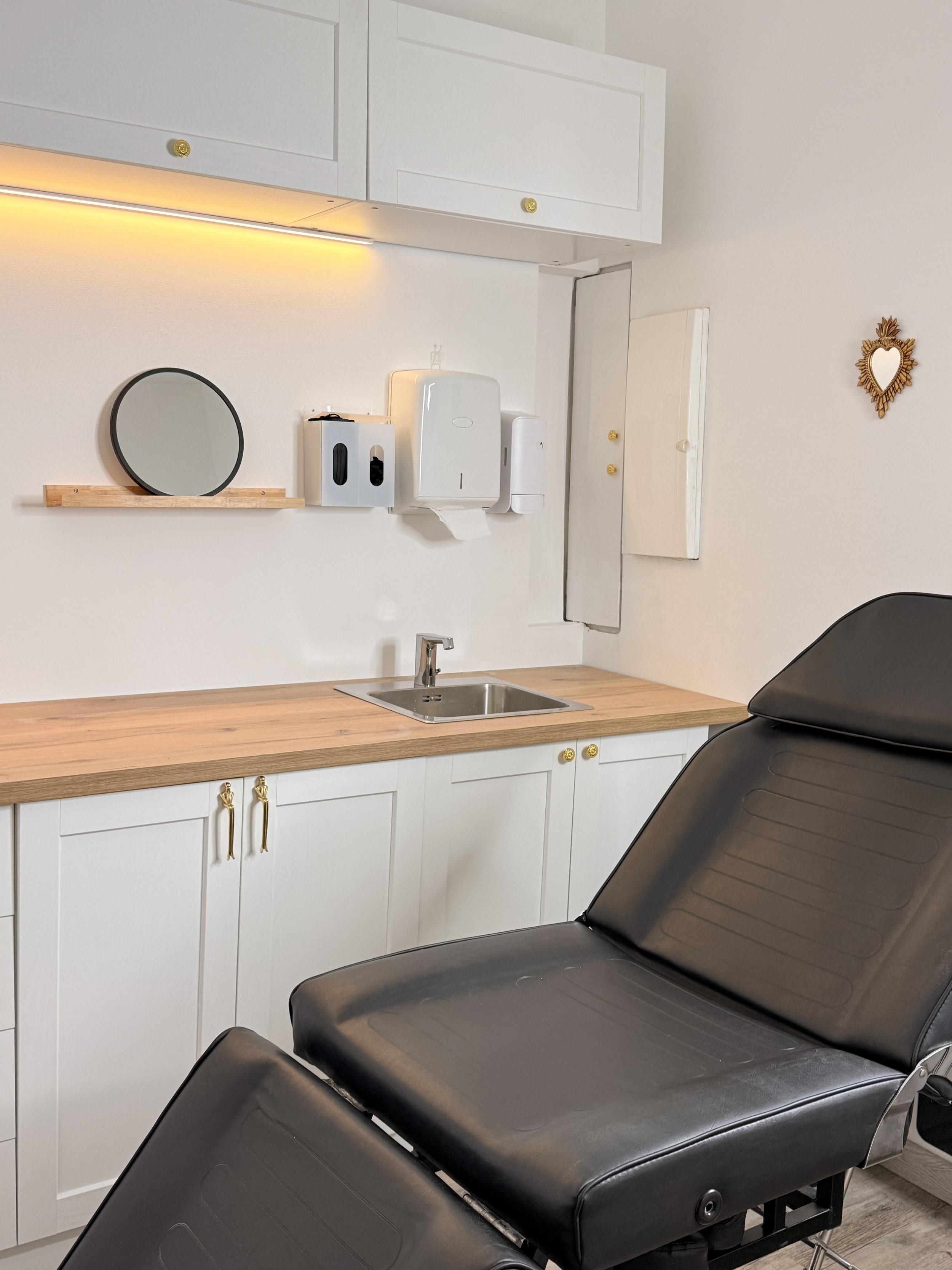 Salle d'examen médical avec un fauteuil d'examen noir, un comptoir en bois avec évier, un miroir rond, un distributeur de gel, une armoire murale, et une décoration murale en forme de cœur.