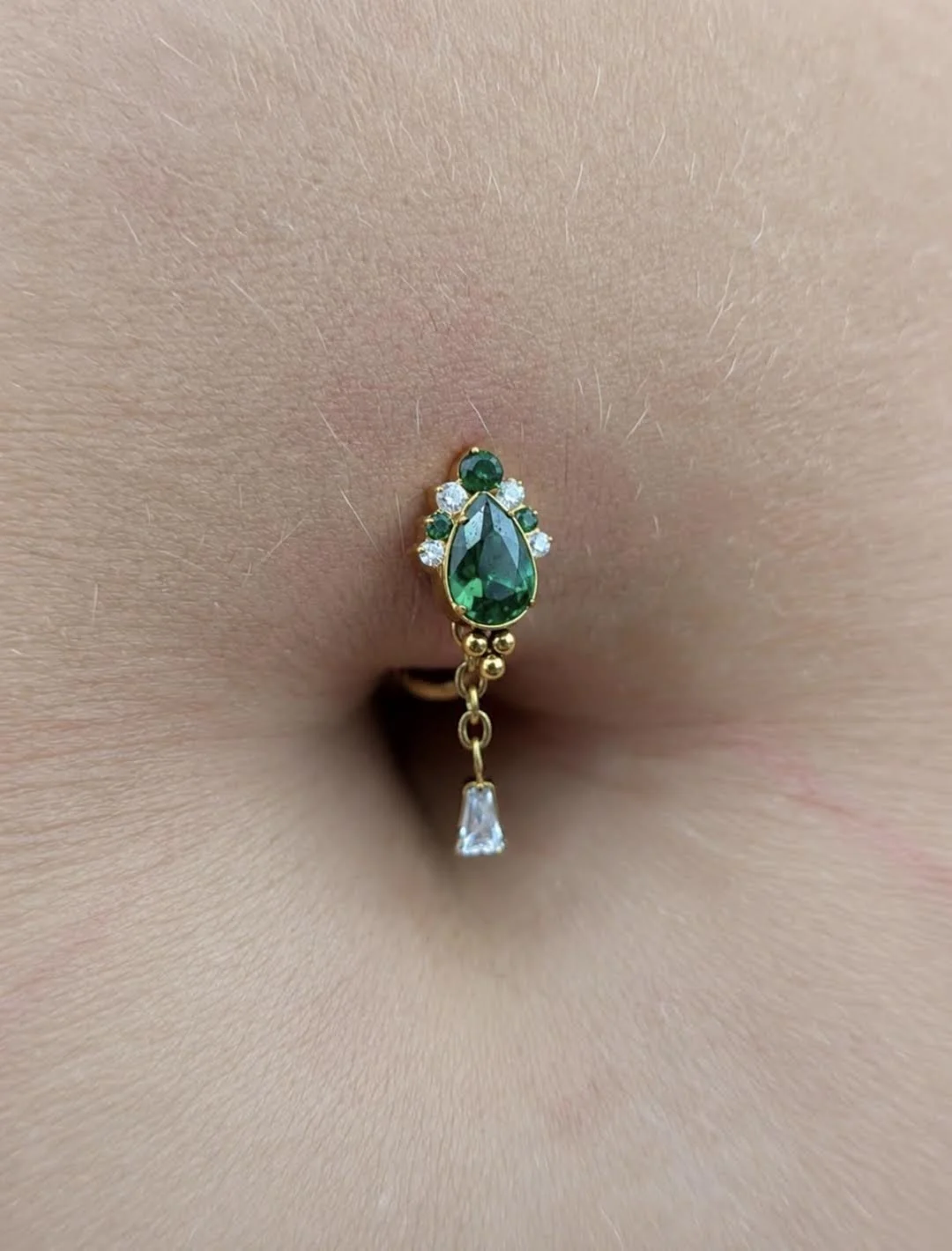 Bijou de piercing au nombril avec un pendentif en forme de goutte en émeraude entourée de petits diamants et une petite cascade avec une pierre en forme de triangle.