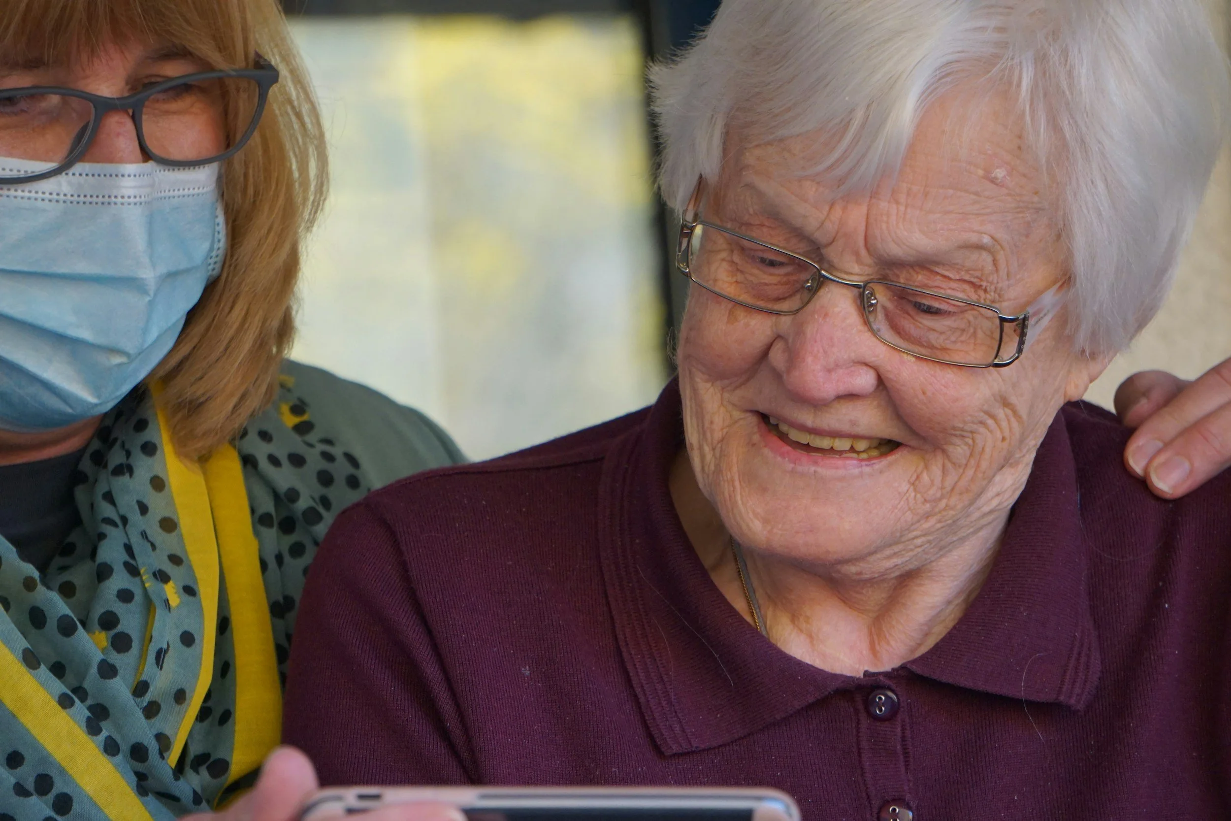 An elderly woman with white hair and glasses smiling while looking at a smartphone, with a caregiver beside her wearing a face mask and glasses, in an indoor setting.