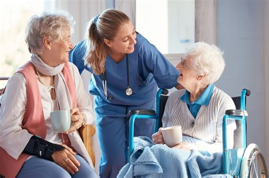 Healthcare worker smiling and talking to elderly woman in wheelchair, with another woman holding a mug nearby.