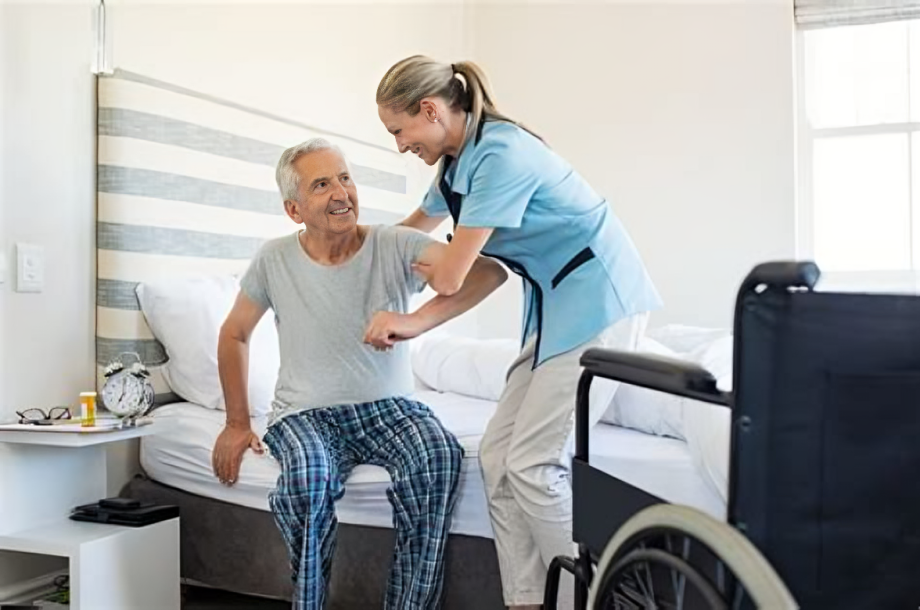 A young female nurse assisting an elderly man with mobility, sitting on the edge of a bed in a hospital room with a wheelchair nearby.