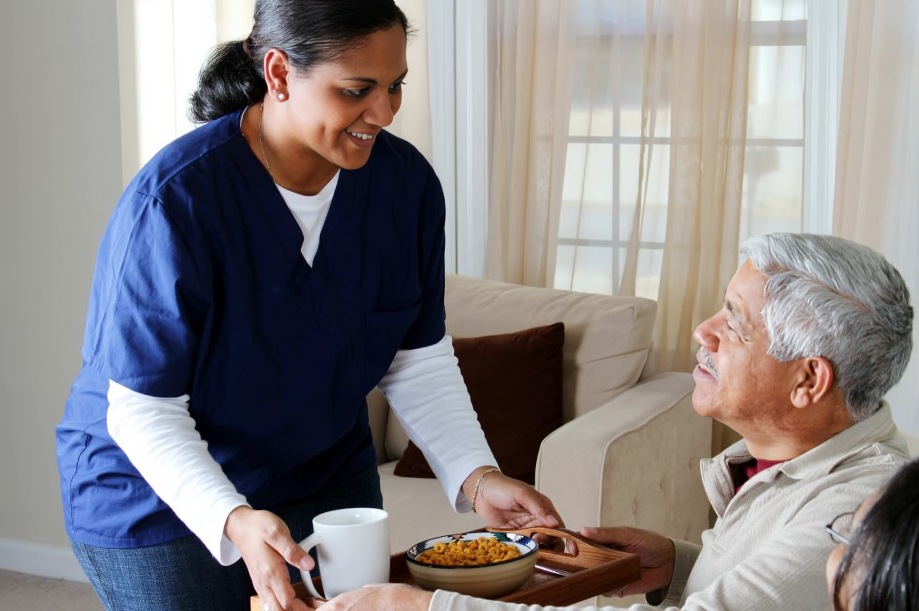 A woman in a blue nurse uniform serves food to an elderly man sitting on a sofa in a well-lit room.