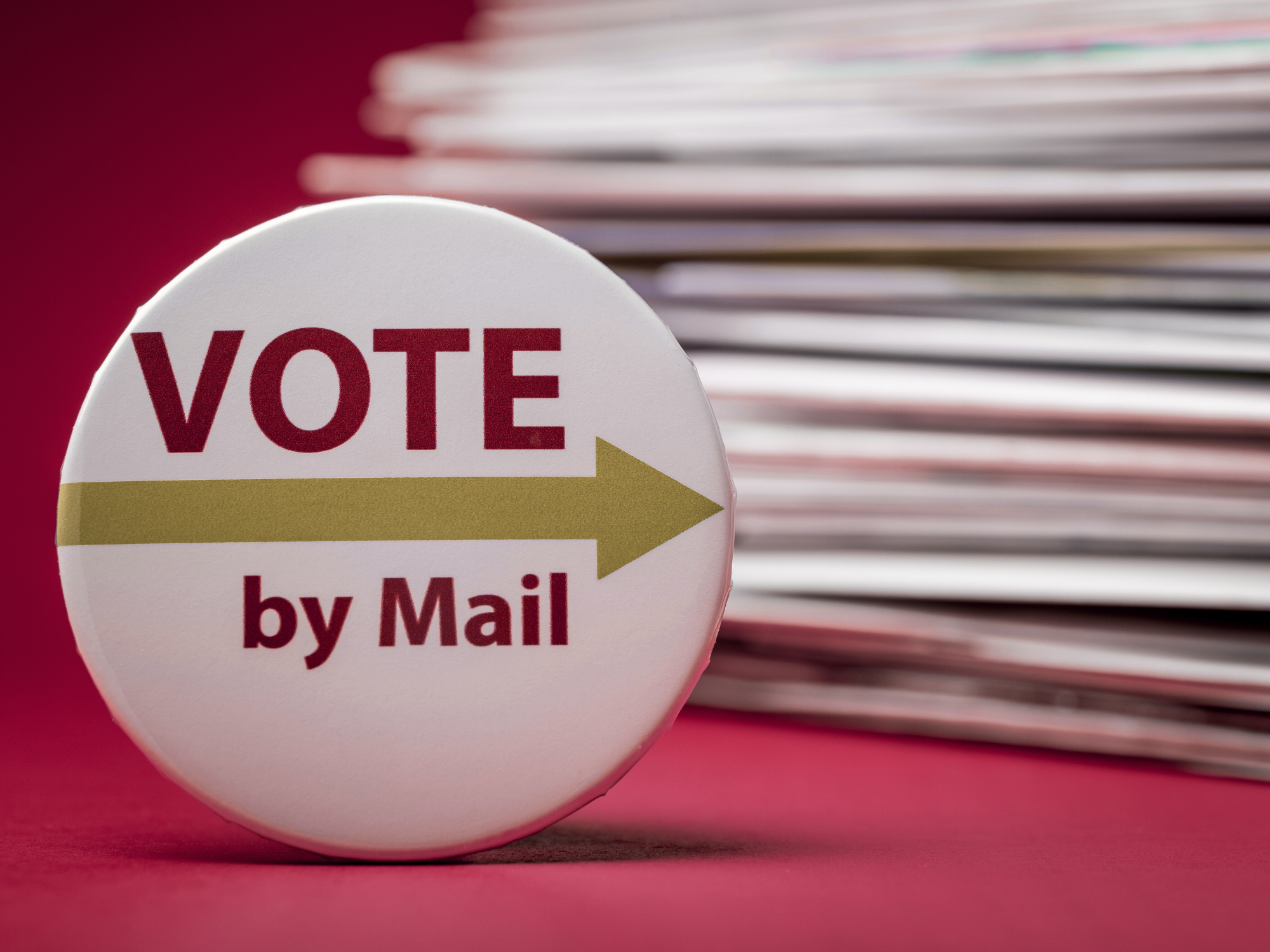 A round button with the words 'Vote by Mail' and an arrow pointing to the right, against a background of stacked paper or mail documents.