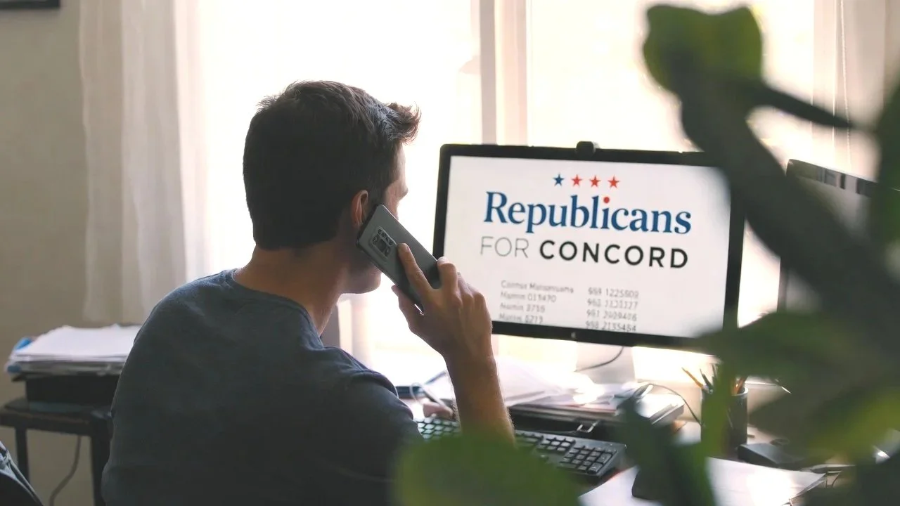 A person sitting at a desk, talking on a phone, with a computer monitor displaying 'Republicans for Concord' and some contact information.