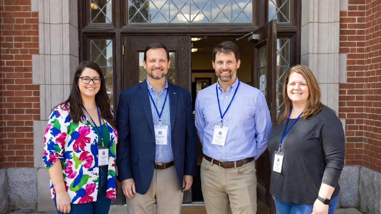 Four people standing in front of a brick building with wooden doors, smiling for the camera, wearing name badges on lanyards.