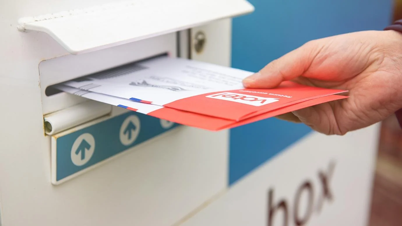 Person inserting a red ballot into a white ballot drop box