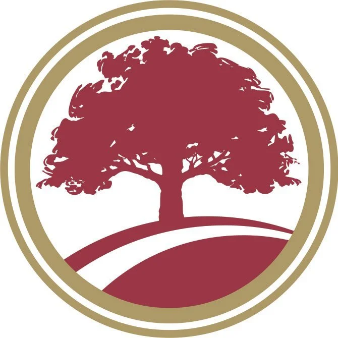 A stylized logo of a large tree with a wide canopy in burgundy, situated on a hill with curved lines, enclosed in a gold and white circular border.