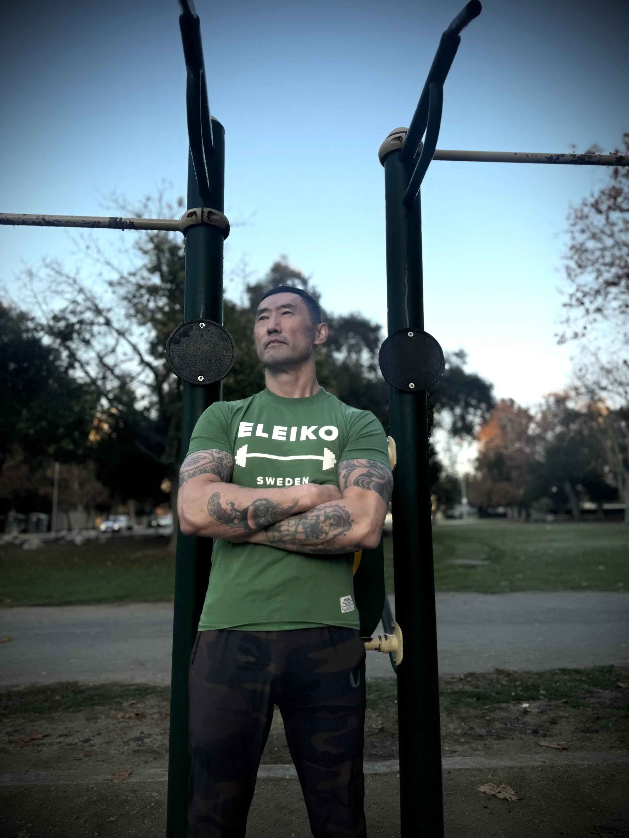 A man with tattooed arms crosses his arms and stands outdoors in front of pull-up bars at dusk.