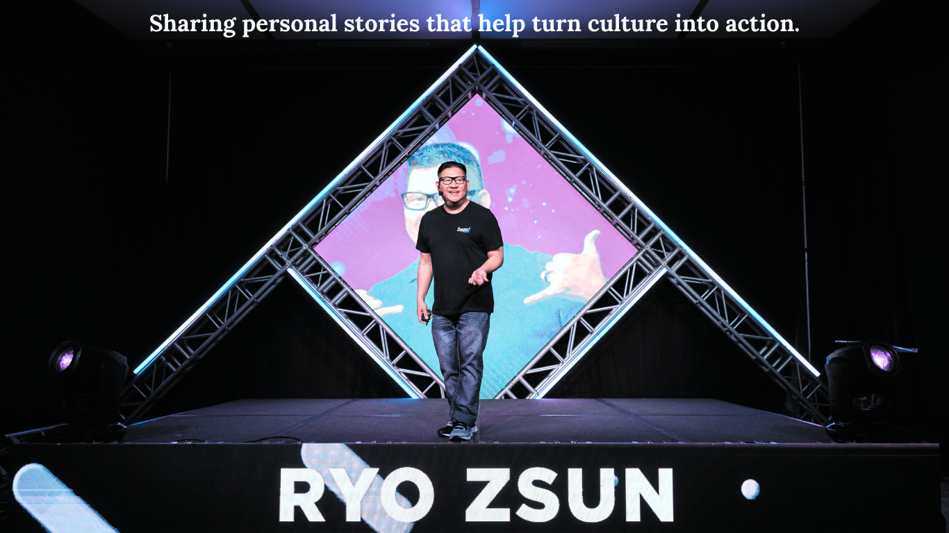 Ryo Zsun smiling on stage at a conference, wearing a black T-shirt and jeans, with his name displayed behind him. The stage features a lit geometric design, and the caption above reads, “Sharing personal stories that help turn culture into action.