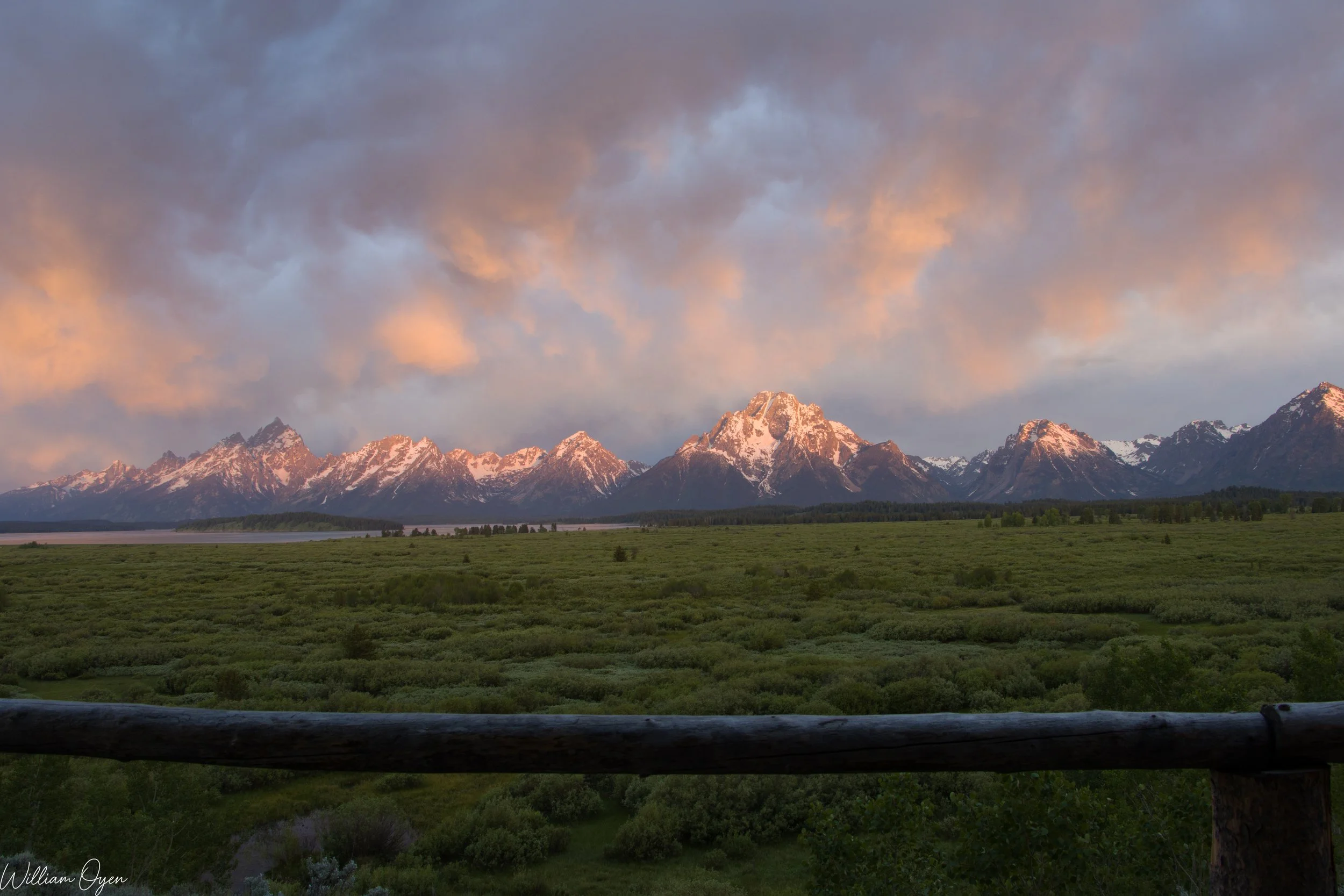 Scenic view of snow-capped mountains at sunset with cloudy sky, green valley in foreground, and a wooden fence in the lower part of the image.