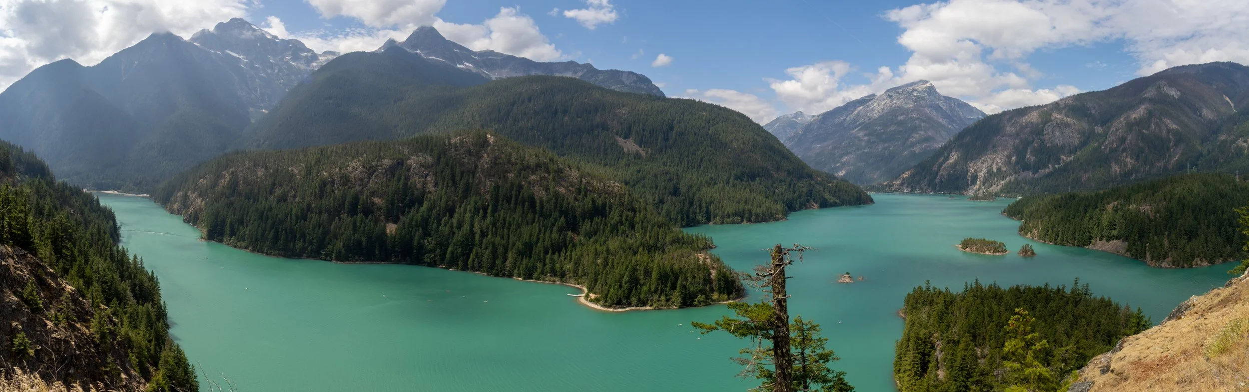 Scenic landscape of a mountain range with a turquoise lake surrounded by evergreen trees and rugged peaks under a partly cloudy sky.