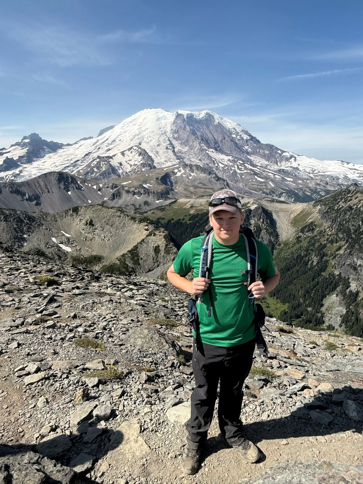 A person in a green shirt and hiking gear standing on rocky terrain with a snow-capped mountain in the background.