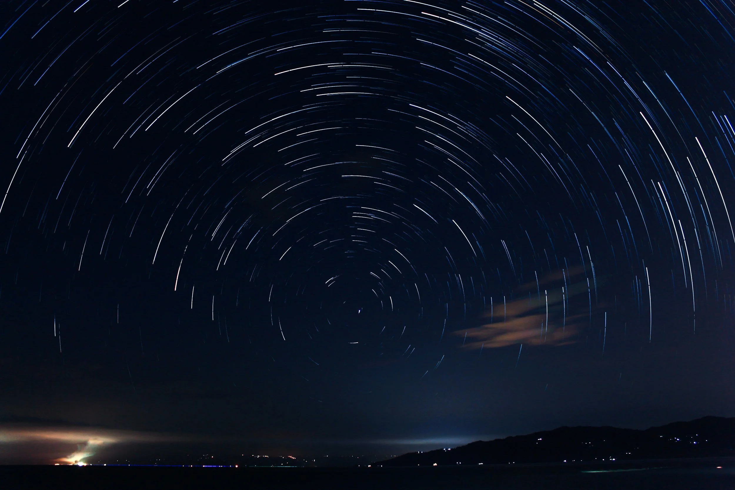 Long exposure photo of star trails over a dark landscape with hills, clouds, and distant lights in the night sky.