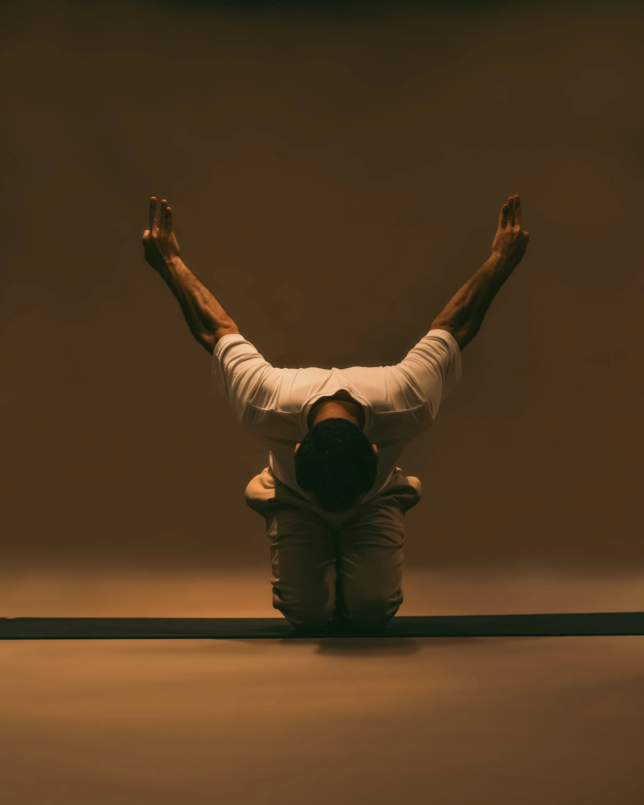 A man in white clothes performing a yoga pose, kneeling with arms raised and hands open, against a plain background.