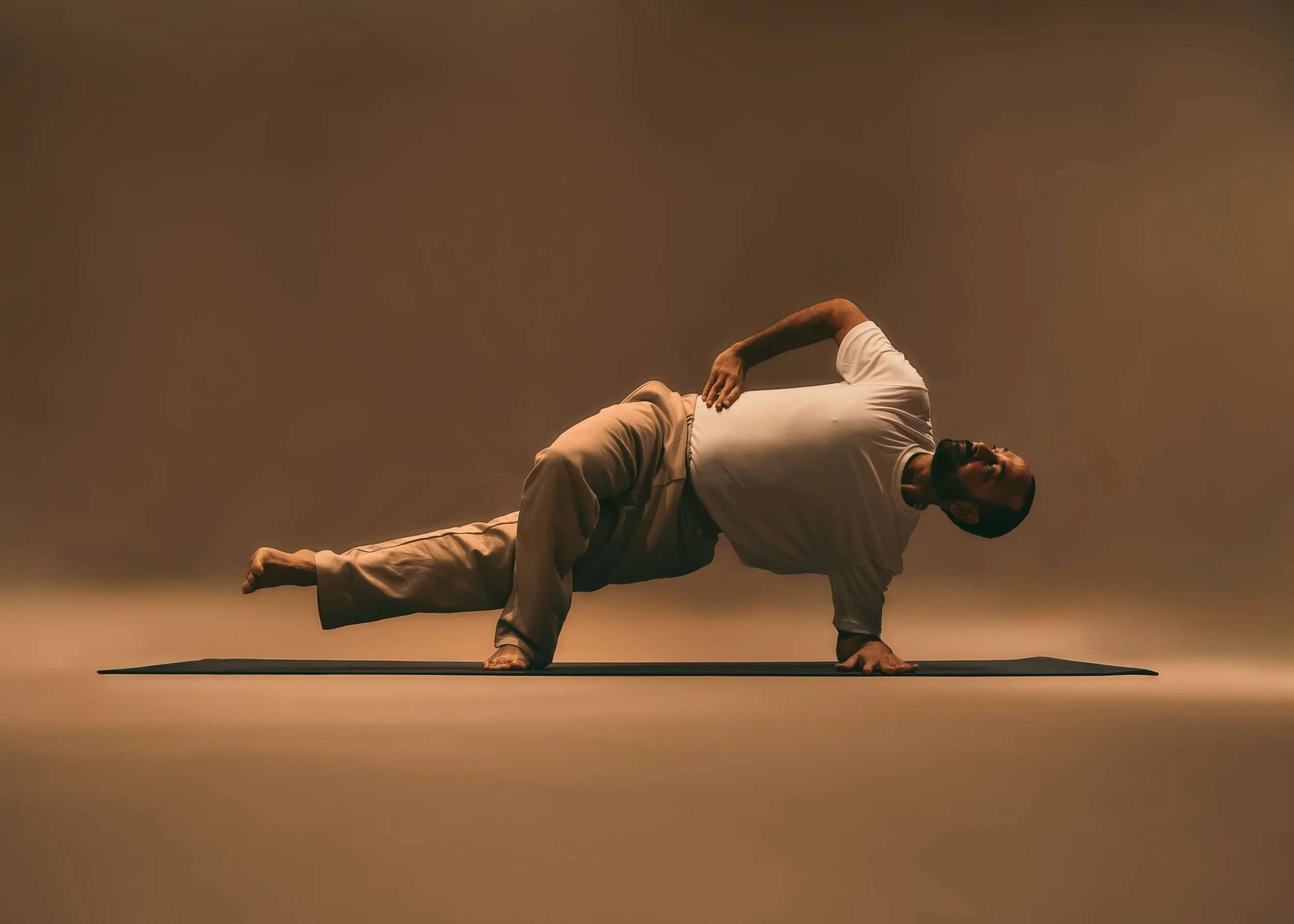 A man performing a one-arm plank pose on a yoga mat in a dimly lit studio.