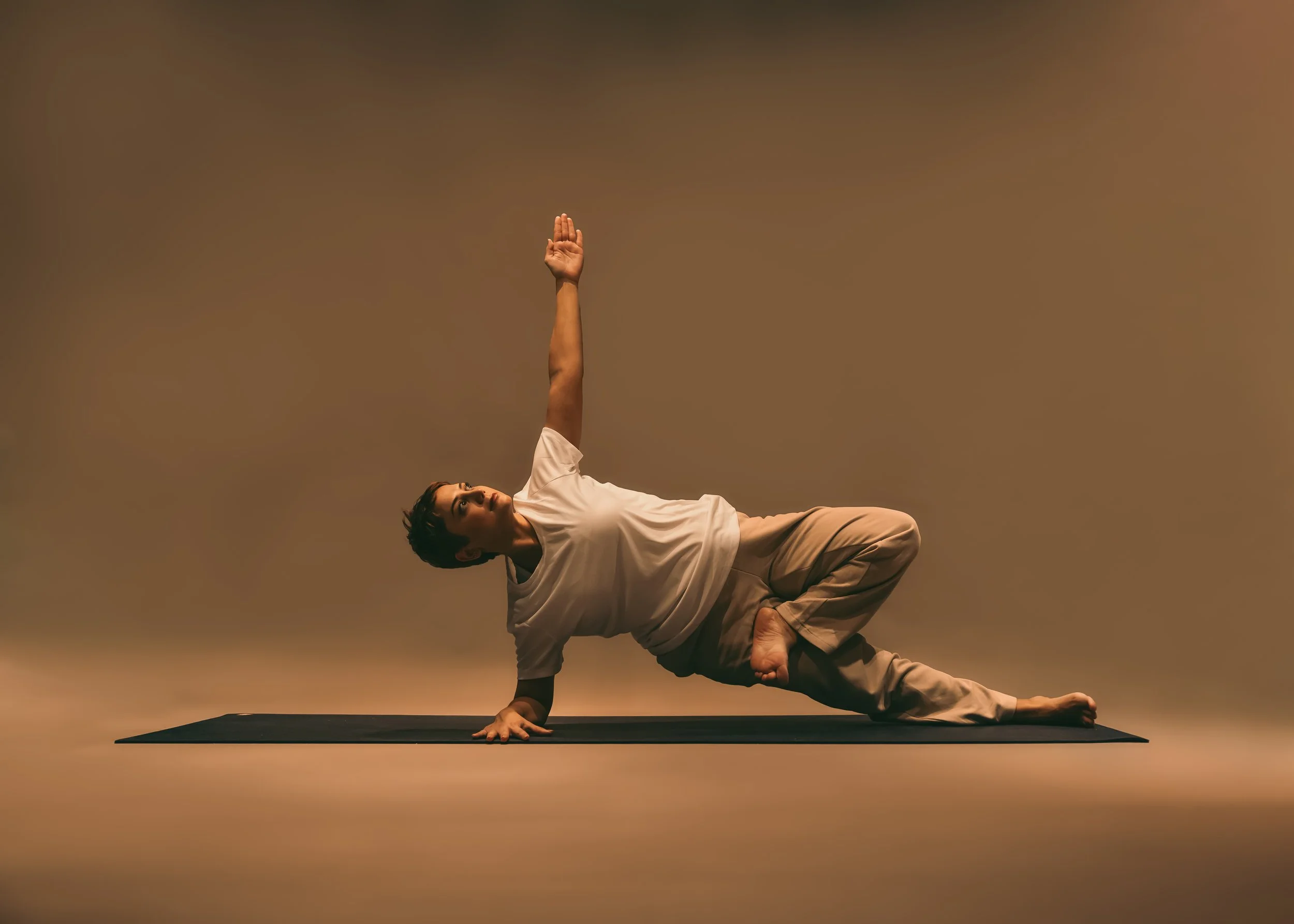 Person practicing yoga on a mat, lying on their side with one arm extended overhead and supported on one hand, in a dimly lit room.