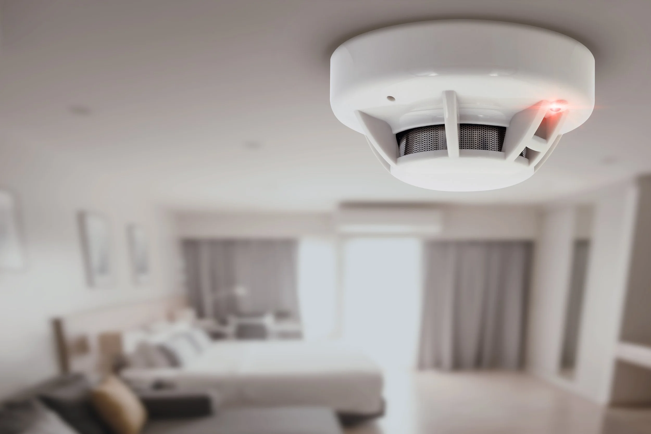 A smoke detector mounted on a white ceiling with a small red LED light turned on, in a blurry living room with a sofa, pillows, a window, and curtains.