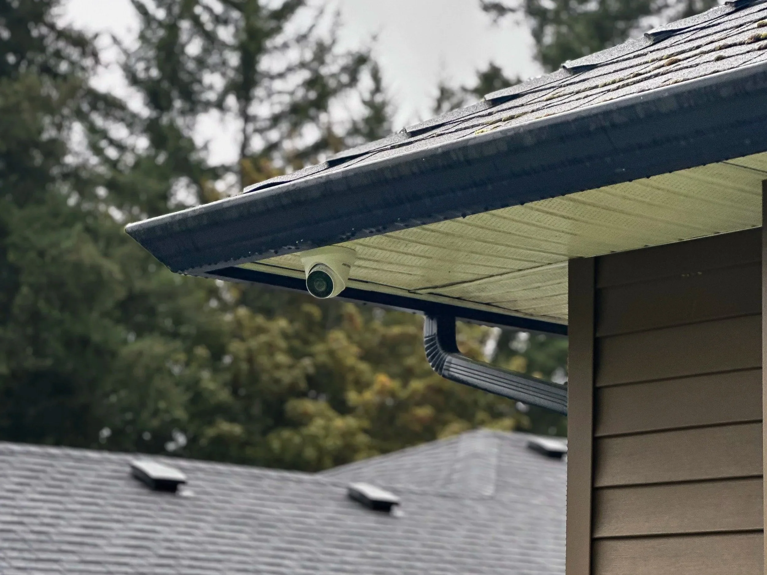 Security camera mounted under the eaves of a house with a brown wall and a gray roof.