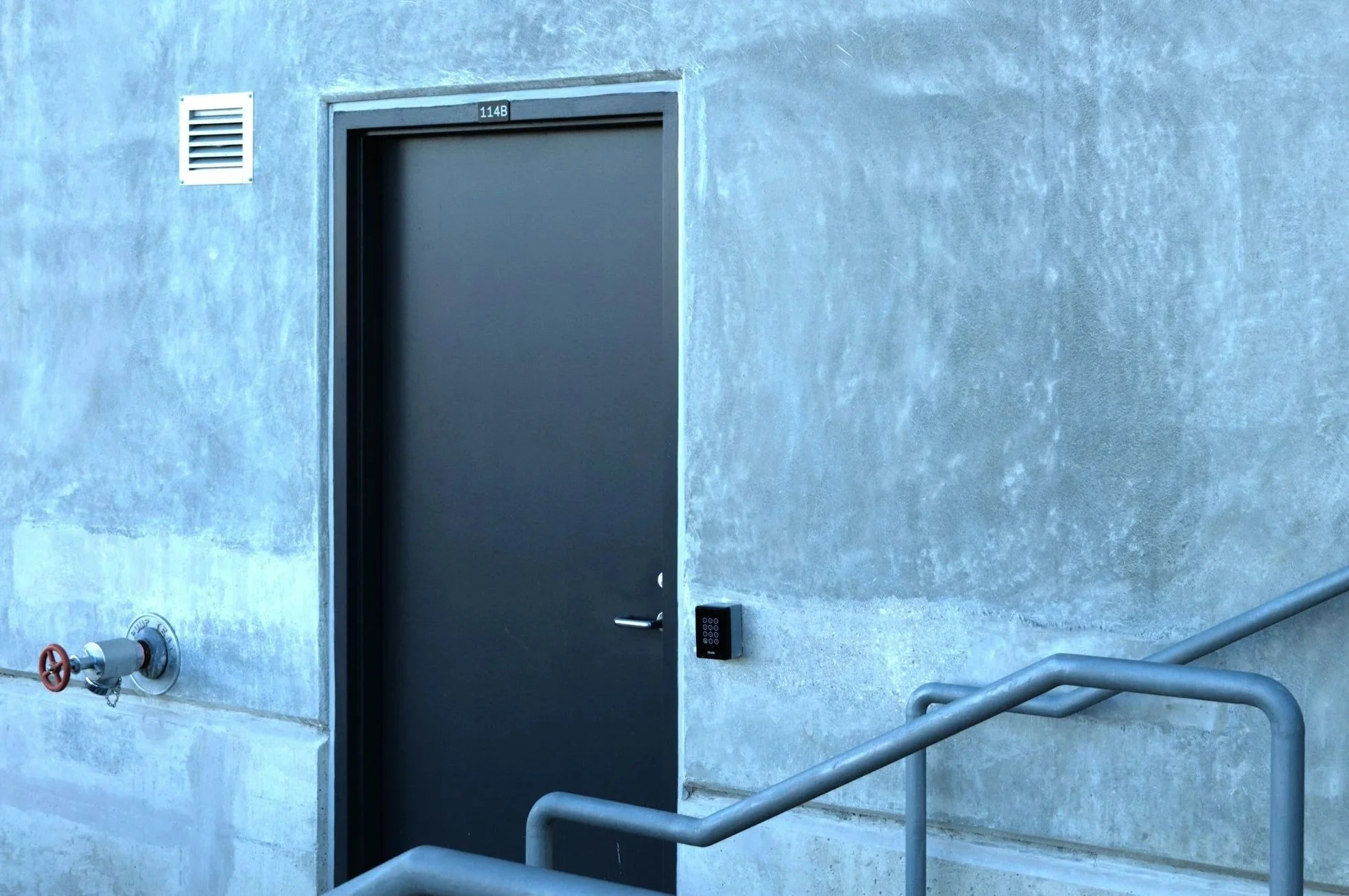 A grey door with a keypad lock on the wall next to it, a red valve on the wall, a white vent, and a metal handrail.