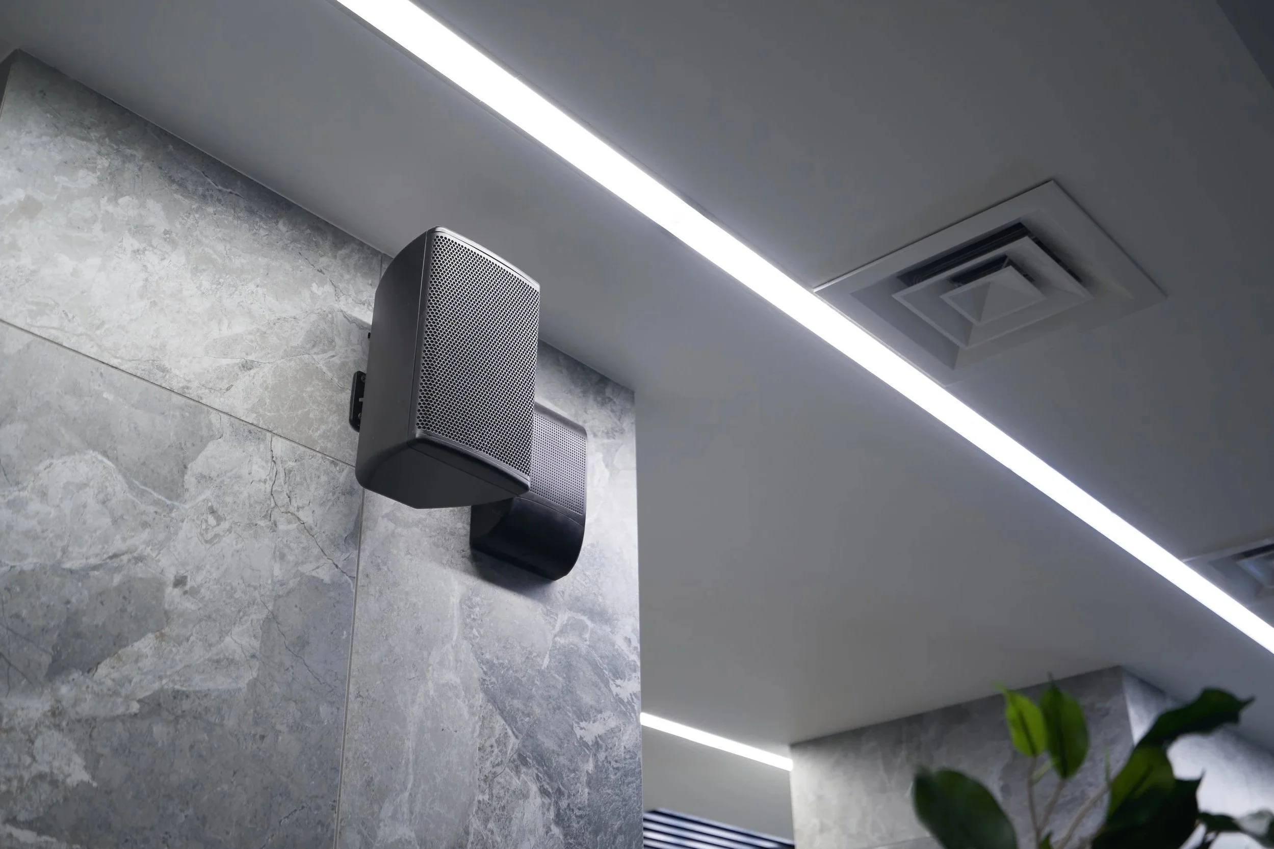 Interior ceiling with light strip, air vent, and mounted speakers on a tiled wall, with a plant in the lower right corner.