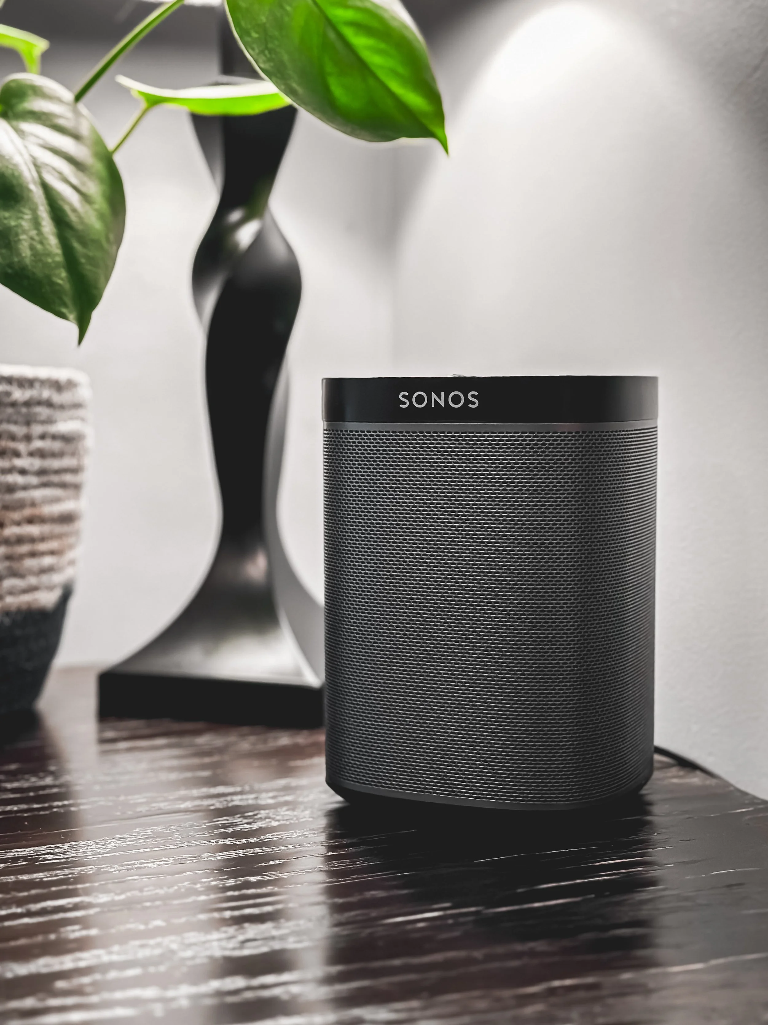 Black Sonos speaker on a dark wooden table with a white wall and decorative objects, including a potted plant and a sculpture, in the background.