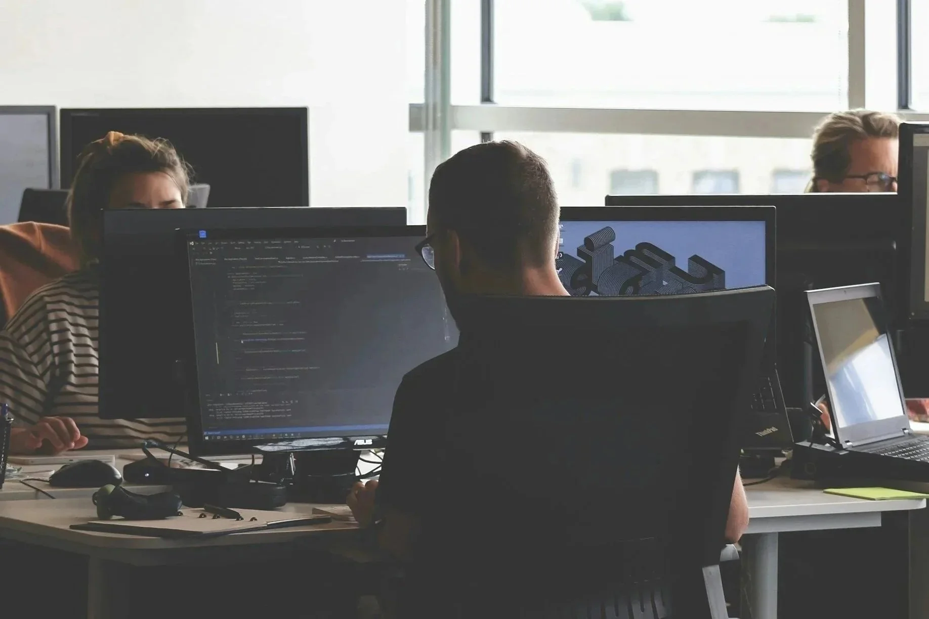 Three people working on computers in an office with large windows. One man in the foreground is looking at a monitor displaying code. Two women are visible, one partially obscured, with computer screens in front of them.