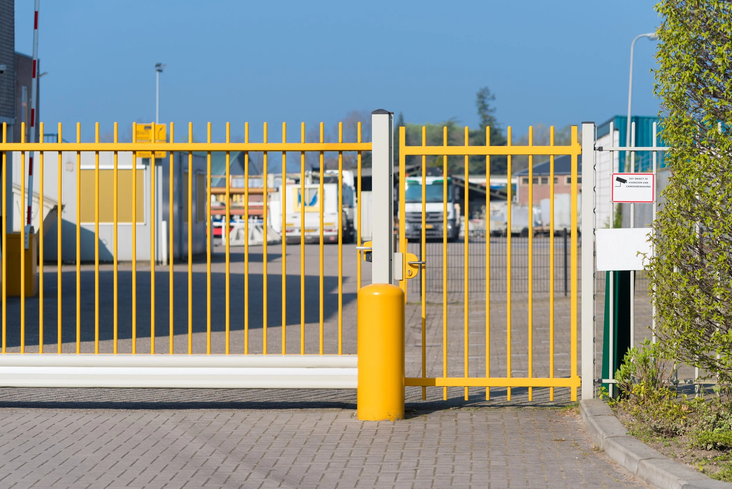 A yellow metal gate with access control lock, separating a parking lot with several trucks parked inside from outside. There is a small sign on the right side of the gate and a large yellow post in front of the gate.