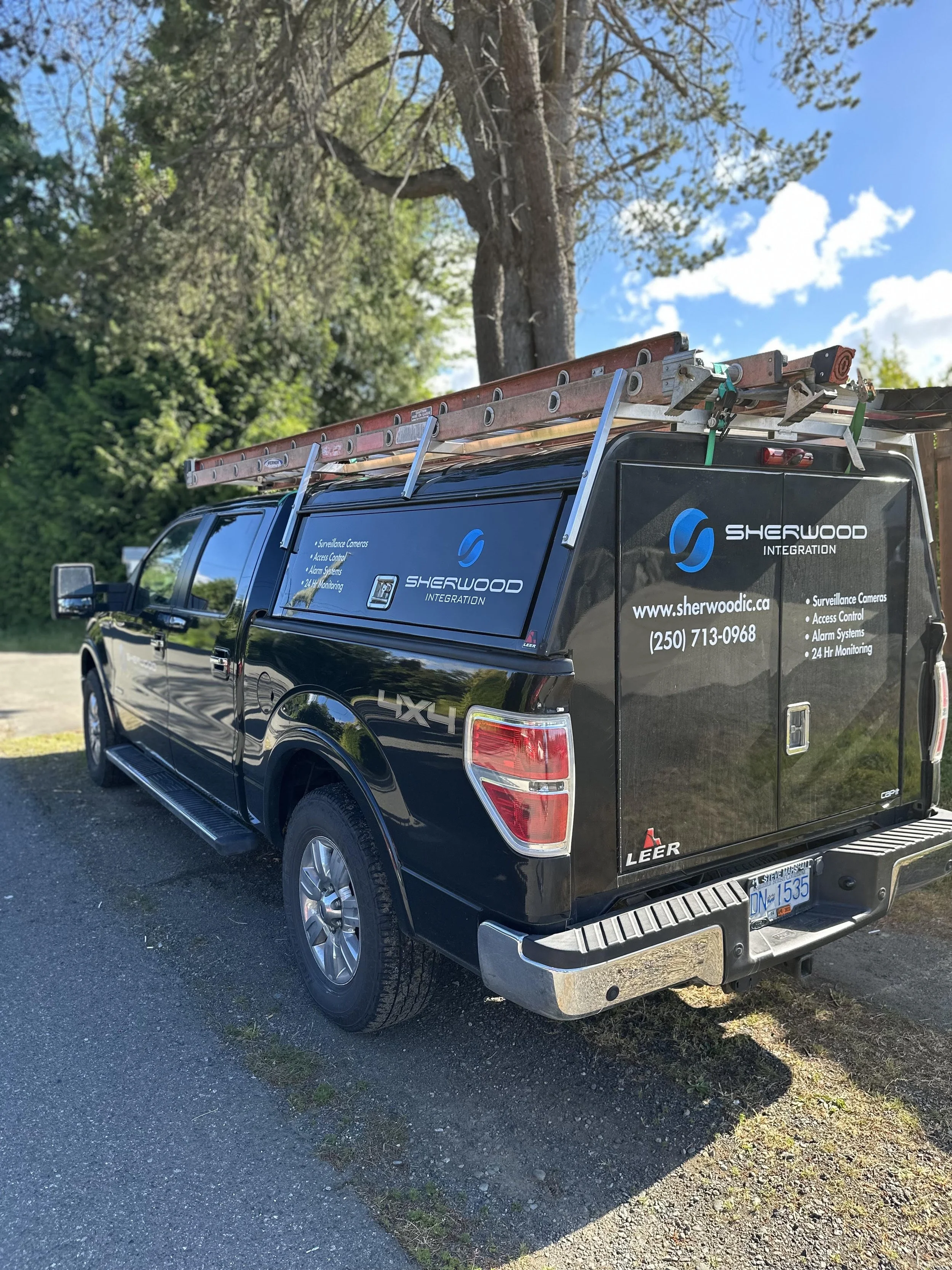Black Sherwood Integration service truck parked outdoors with a ladder on top, surrounded by trees and a partly cloudy sky.