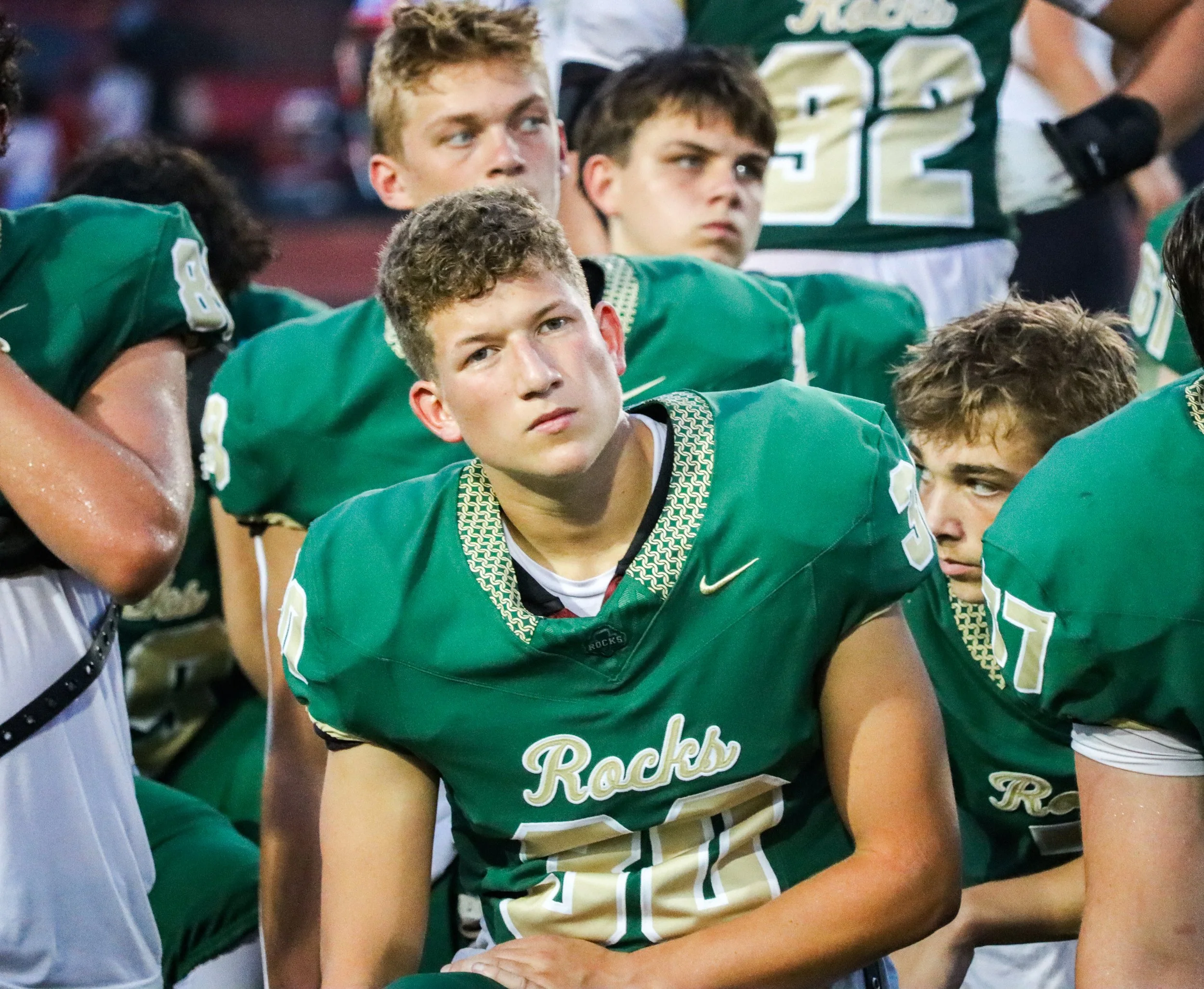 A group of football players in green jerseys, kneeling during a game or practice, with focus on a young player in the foreground.