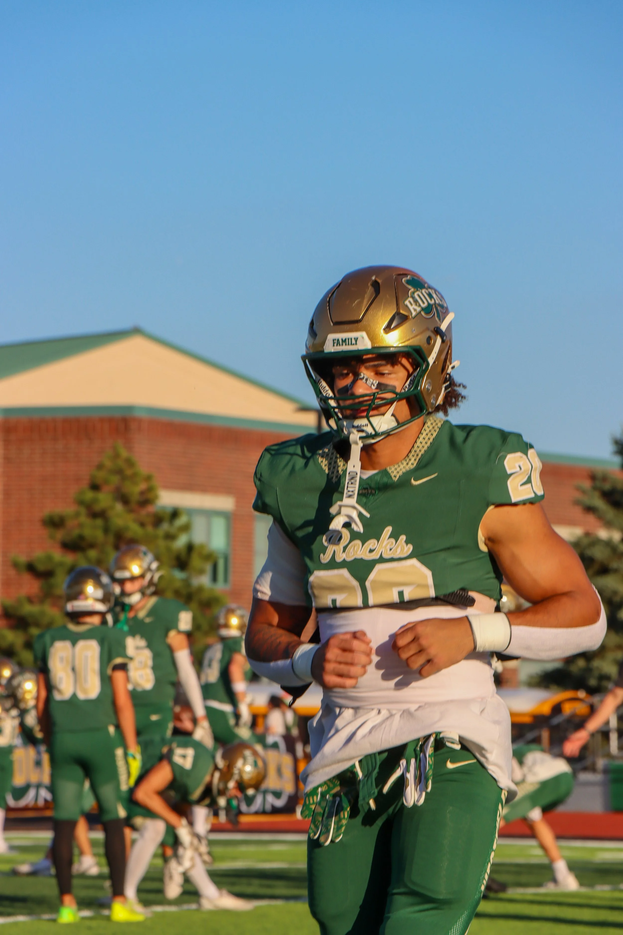 A football player wearing a green and white uniform with a gold helmet, participating in a game on a football field with teammates in the background.