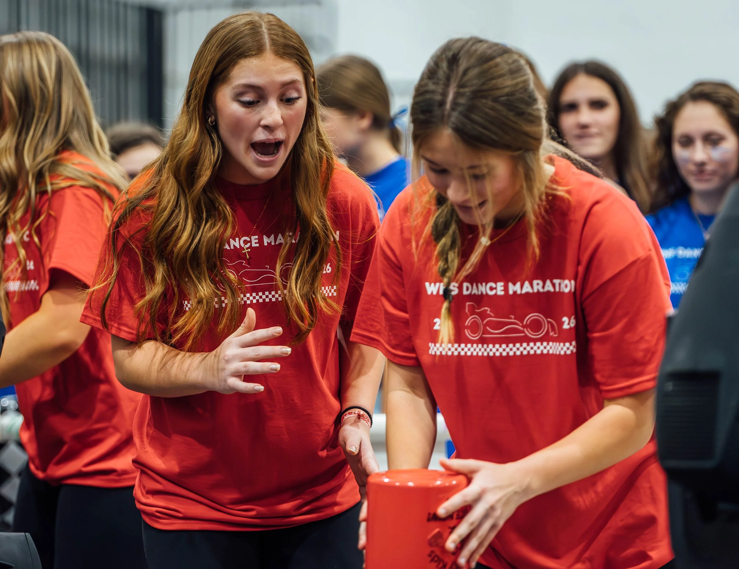 A group of women wearing red shirts with racing-related text and graphics, gathered around a container, likely preparing for or participating in a race event.
