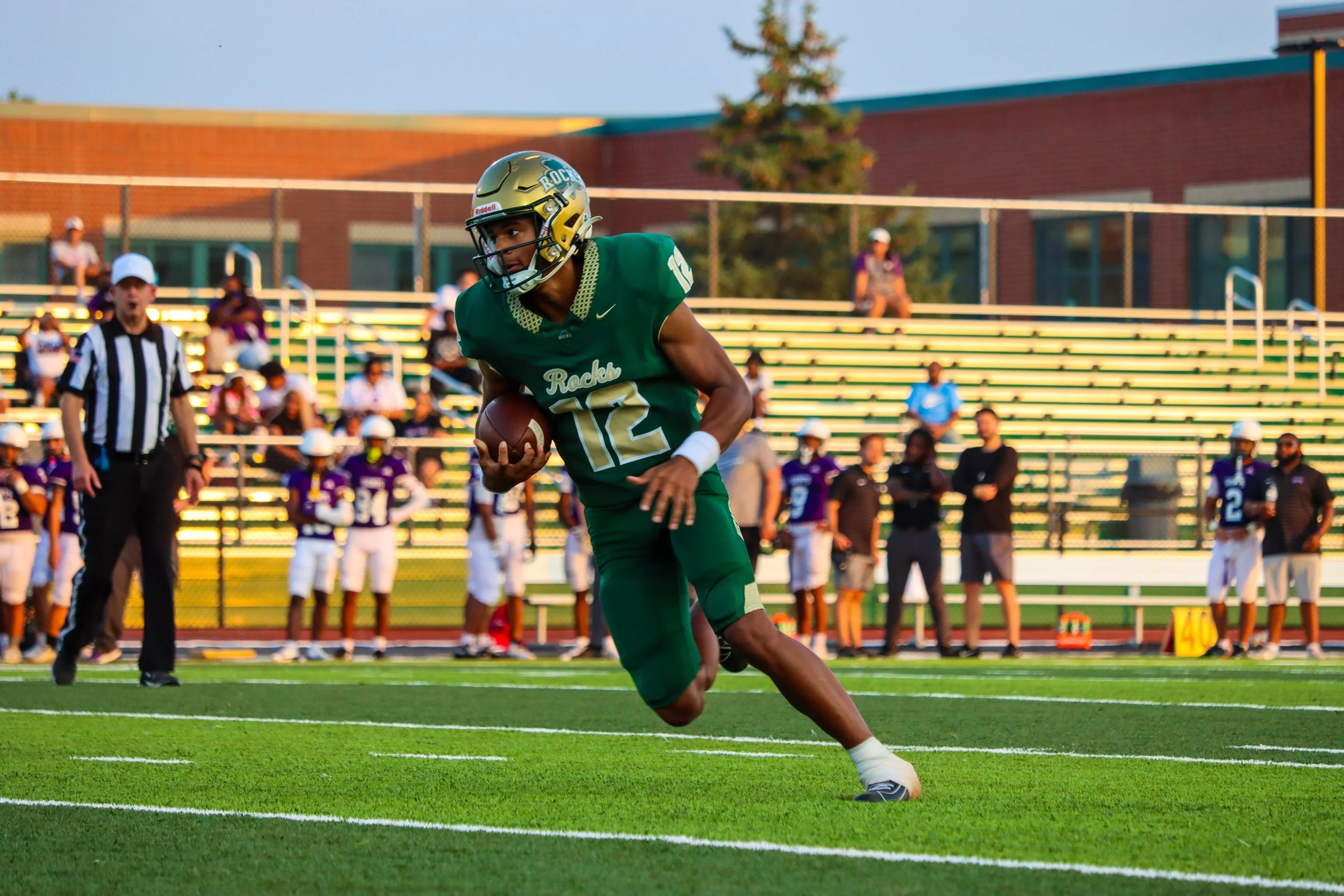 A football player in a green uniform with the number 12 runs with the ball on the field, with spectators and teammates in the background.