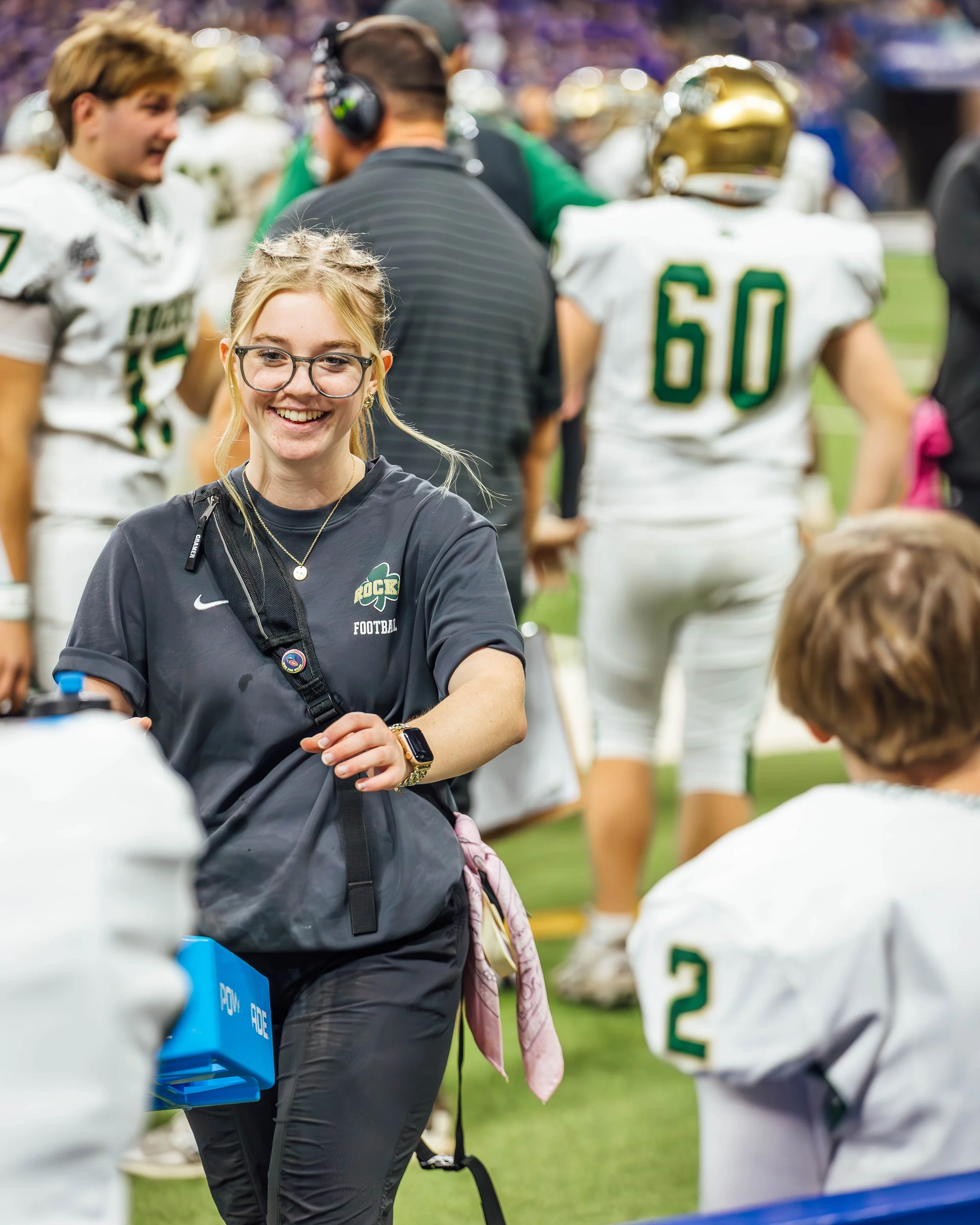 Smiling young woman with glasses and blonde hair, wearing a black sports jacket with a logo and a black backpack, at a football game. She is standing on a field with players and others in the background.