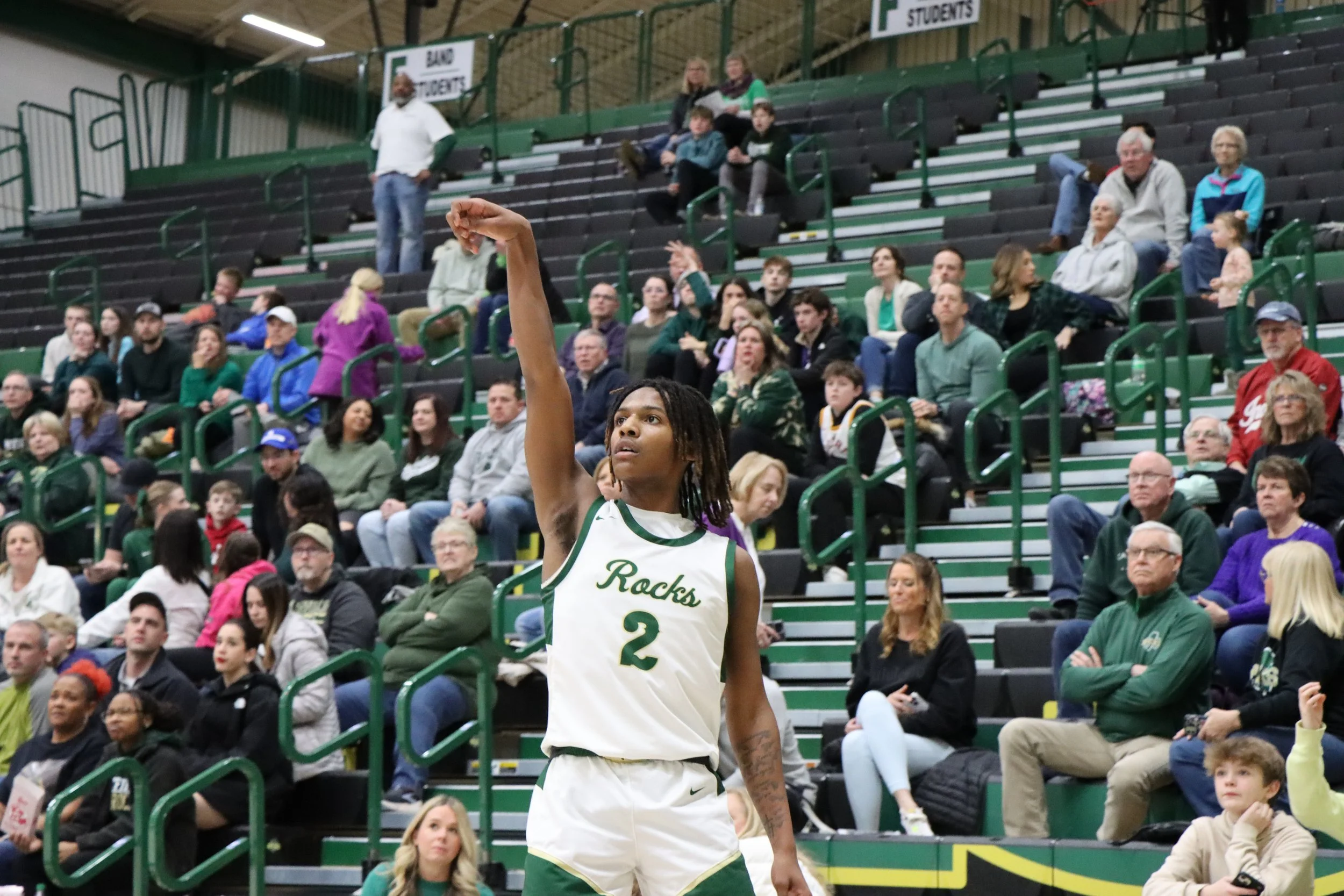 Basketball player in a white jersey with green accents and the words 'Rocks' and the number 2, standing on a gymnasium court, with audience seated in green bleachers around him.