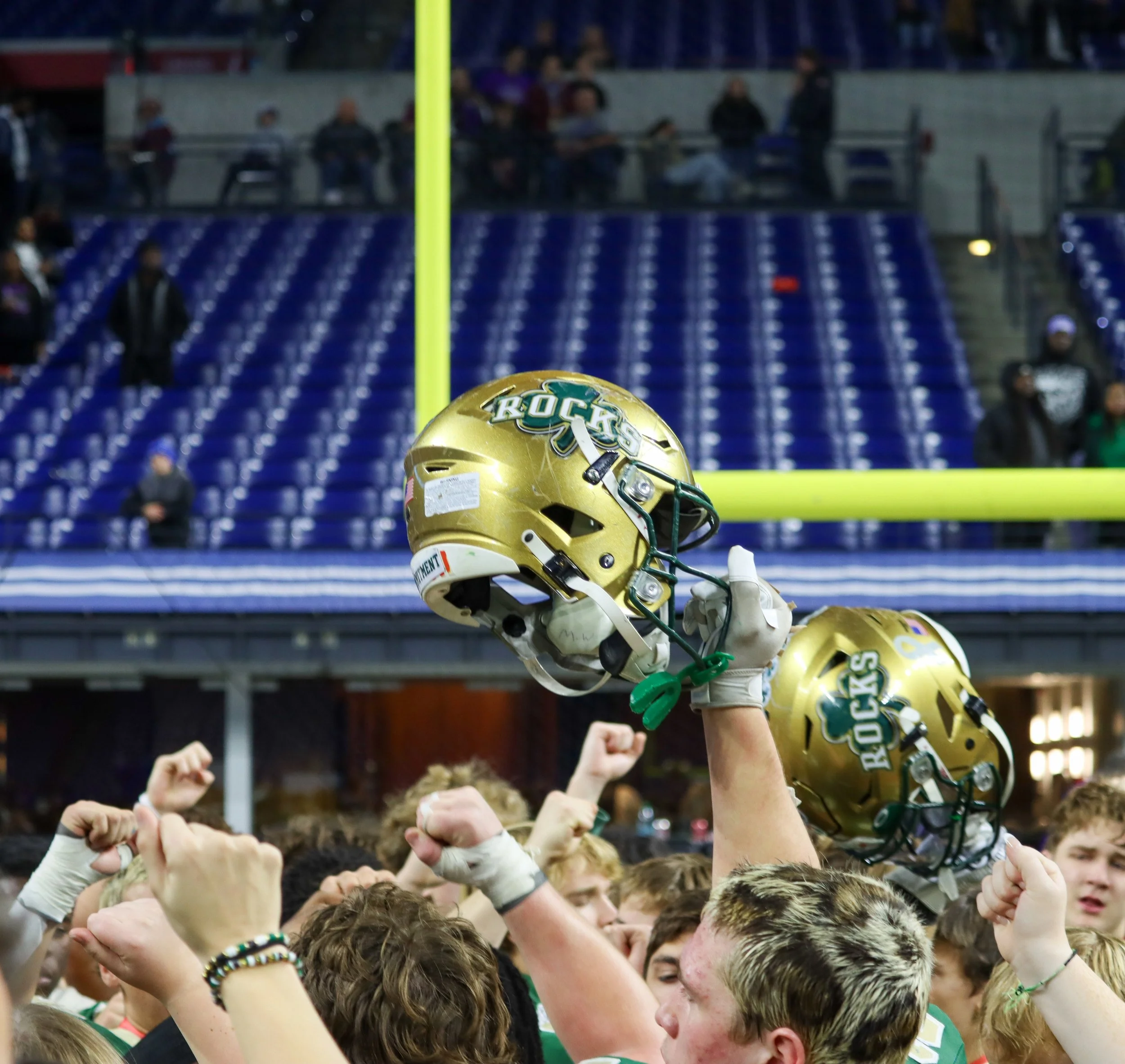 A gold football helmet with the word 'ROCKS' and a green logo on it, held above a crowd of excited people at a sports stadium, with their fists raised in celebration.
