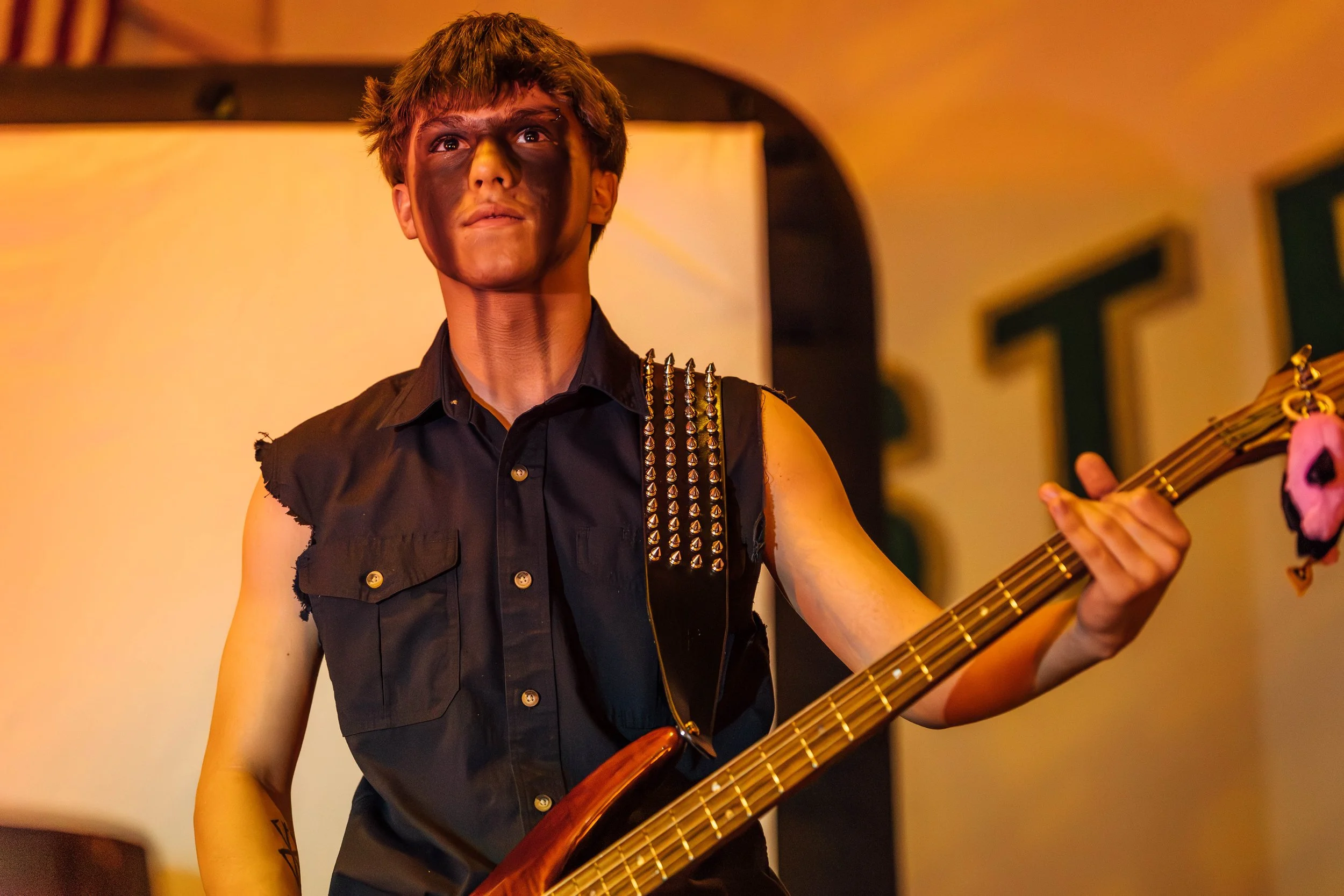 Young woman playing an electric bass guitar with a sleeveless black vest decorated with metal studs, standing in front of a yellow-orange background.
