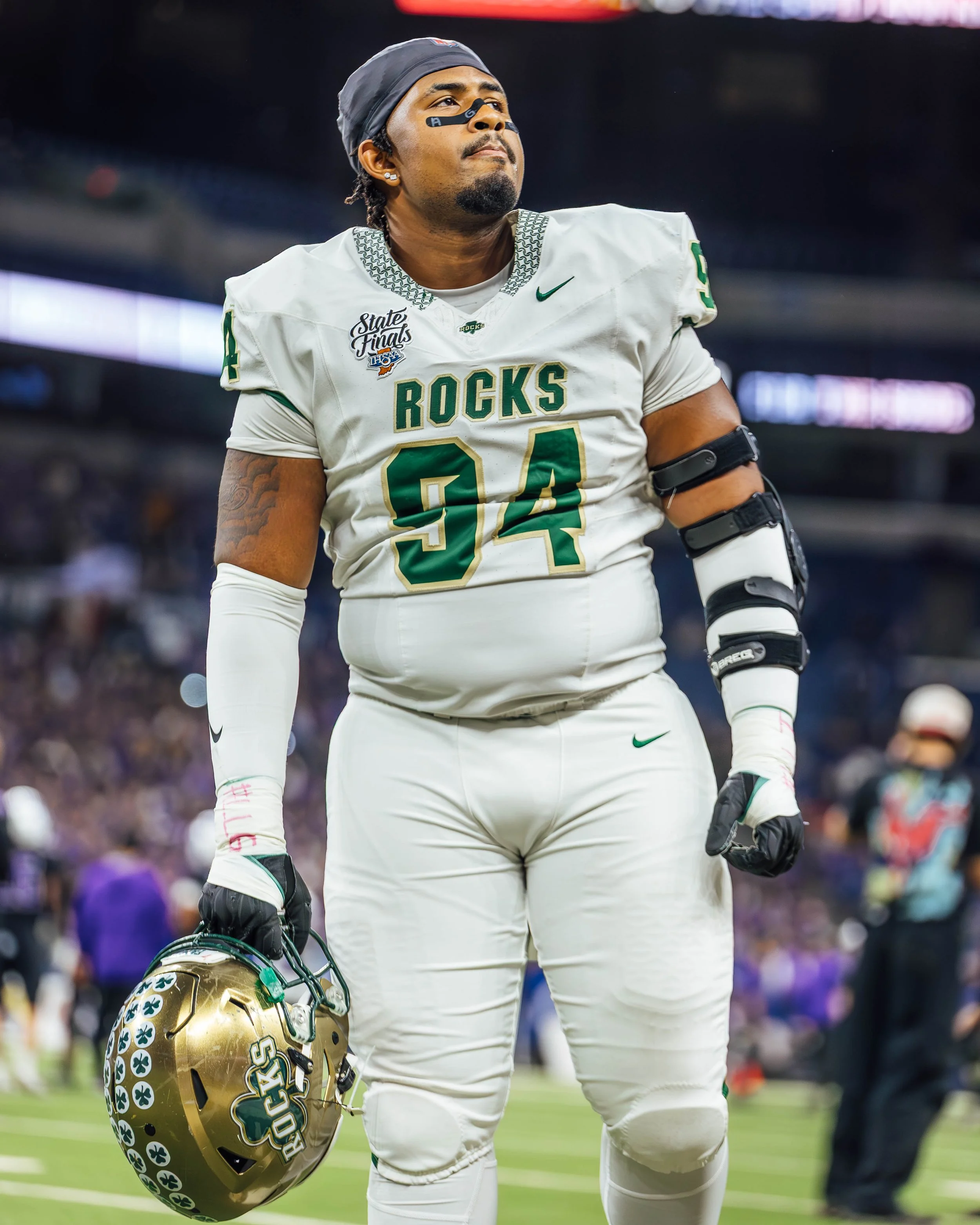 A football player in a white uniform with 'Rocks' and the number 34 on it, standing on a football field holding a helmet with the same logo and a gold face mask, is looking thoughtfully into the distance. He is wearing a black eye shield, earrings, a