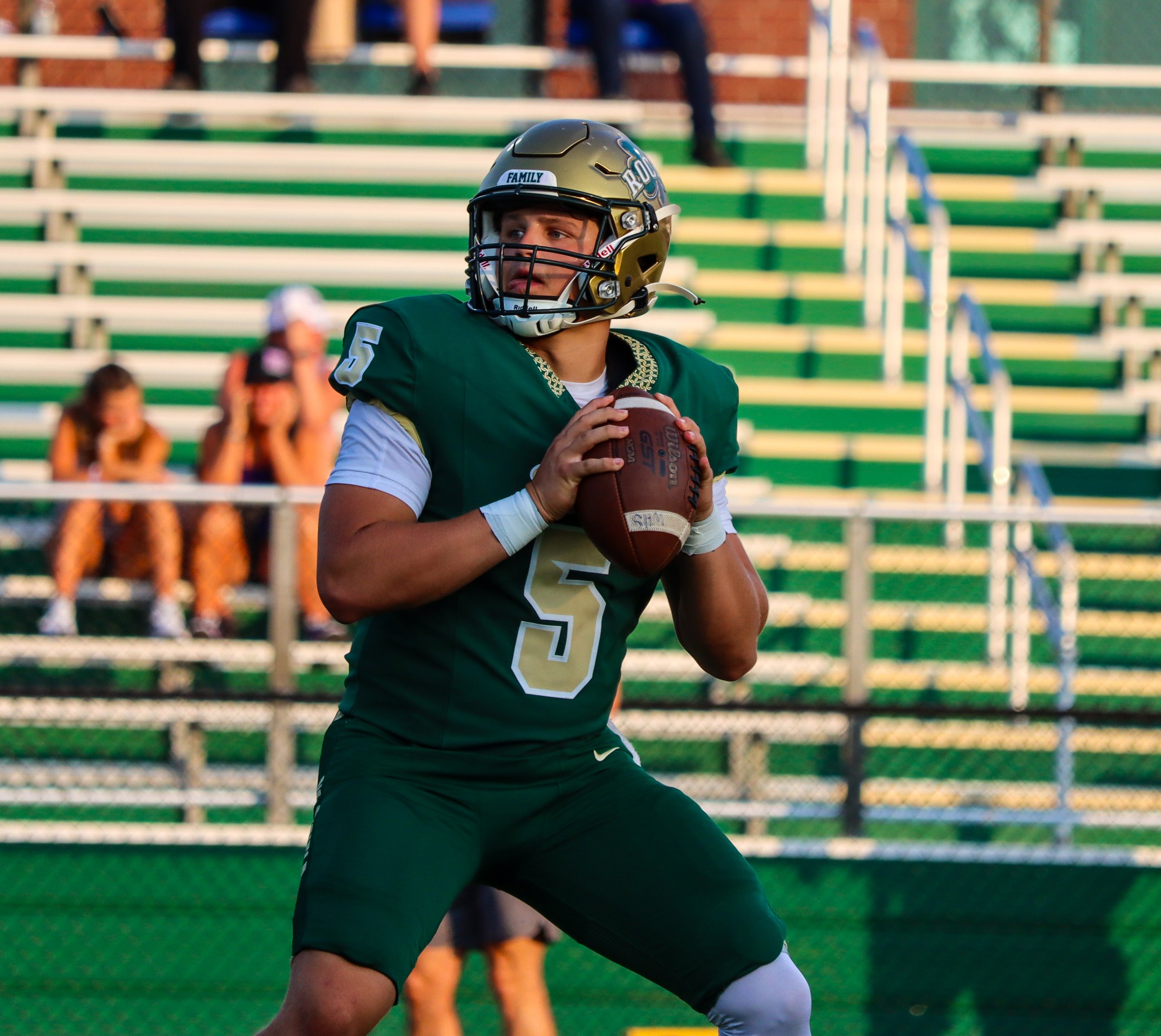 A football player in a green and white uniform, wearing a helmet with a faceguard, prepares to throw a football on the field with empty bleachers in the background.