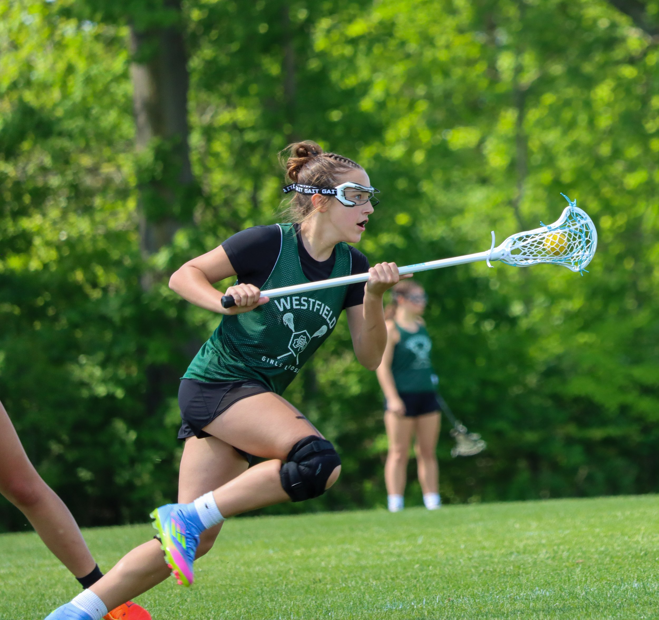 A female lacrosse player in green uniform with black sleeves, wearing protective goggles, is running on a grassy field holding a lacrosse stick with a ball in the netted pocket.