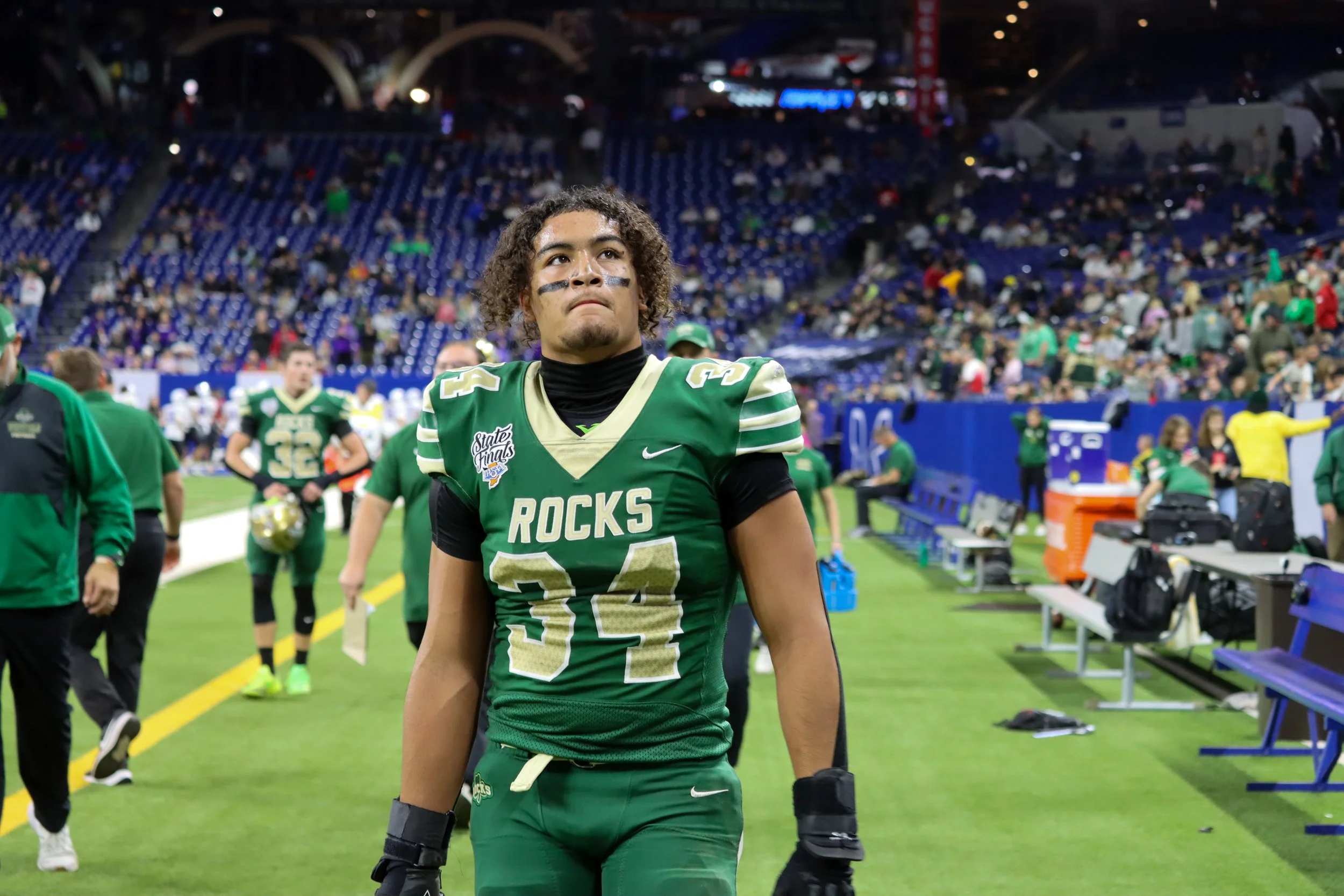 Football player wearing green and gold jersey with the number 24 on it, standing on a football field after a game at night, with empty stadium seats and spectators in the background.