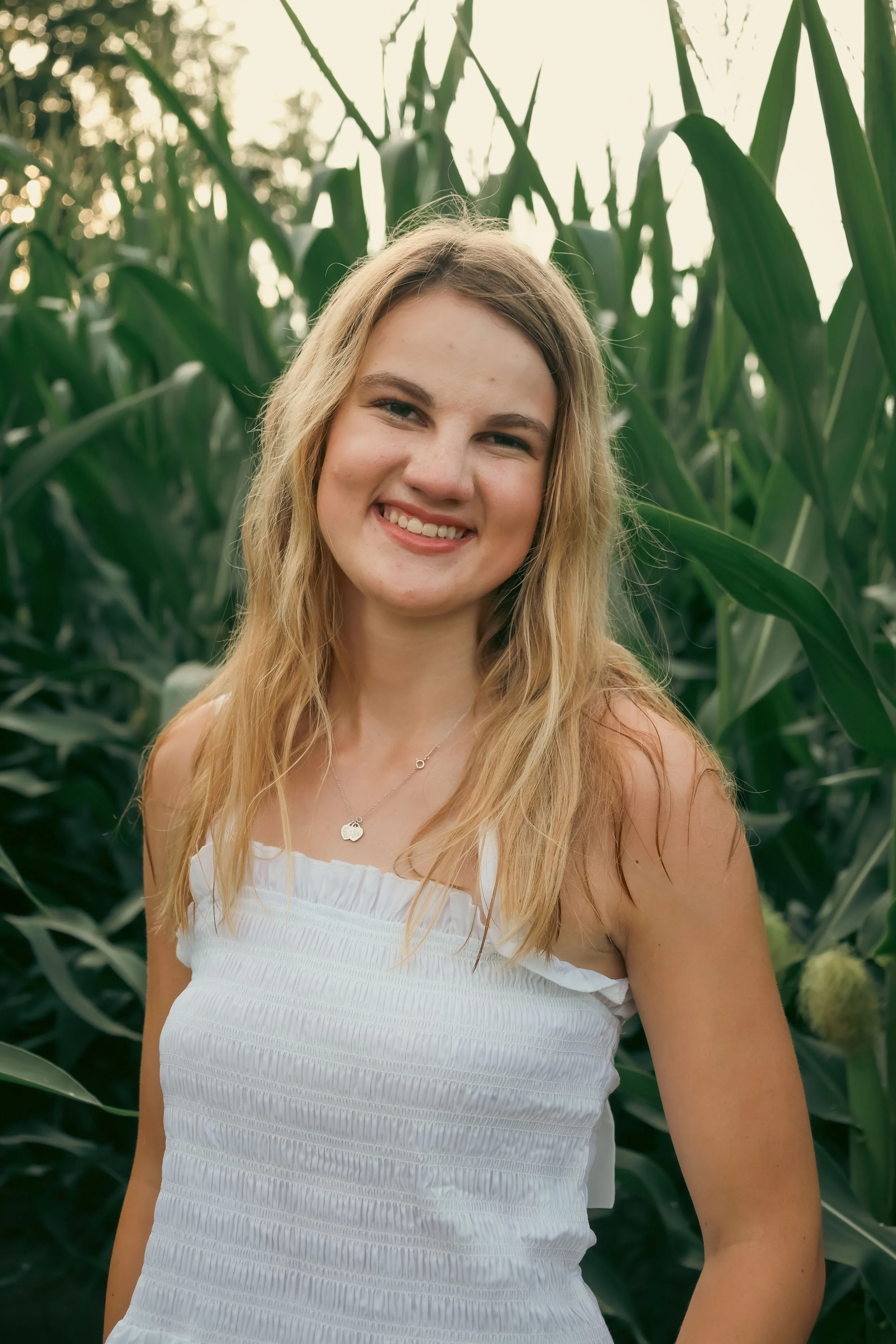 A young woman with long blonde hair smiling in a field of tall green corn plants, wearing a white sleeveless top and a silver necklace.