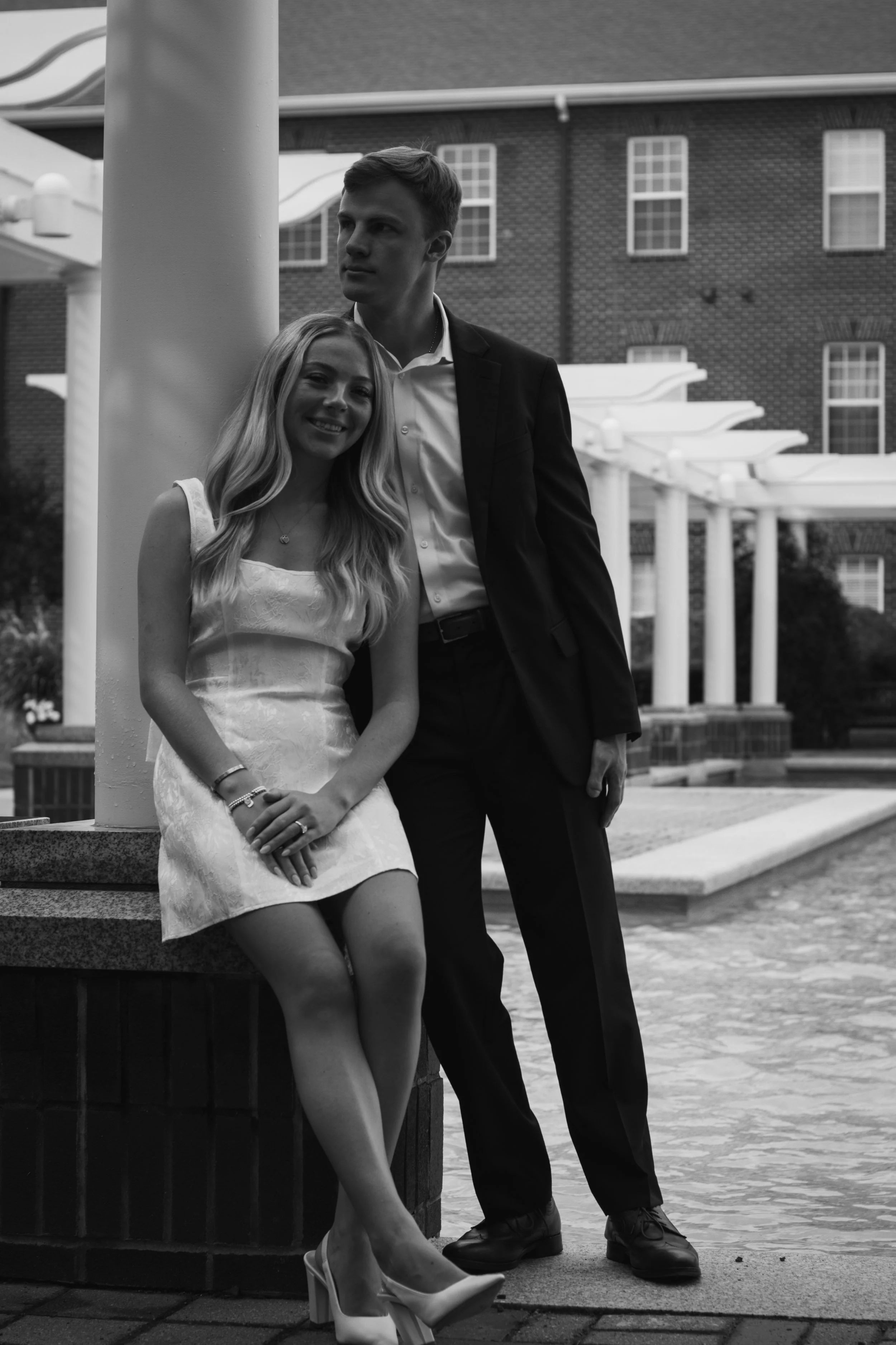 A black and white photo of a young woman in a dress and high heels sitting on a ledge, smiling, with a young man in a suit standing nearby and leaning against a pole. The background features a building with multiple windows and a covered walkway with